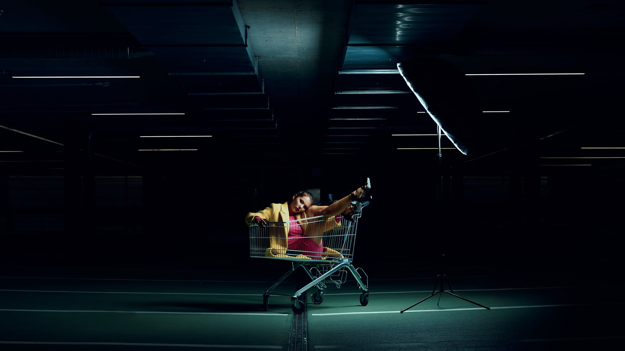 Une personne vêtue de vêtements clairs pose de manière ludique à l'intérieur d'un chariot de supermarché dans un parking faiblement éclairé, avec une jambe levée et une grande lampe de studio à côté d'elle.