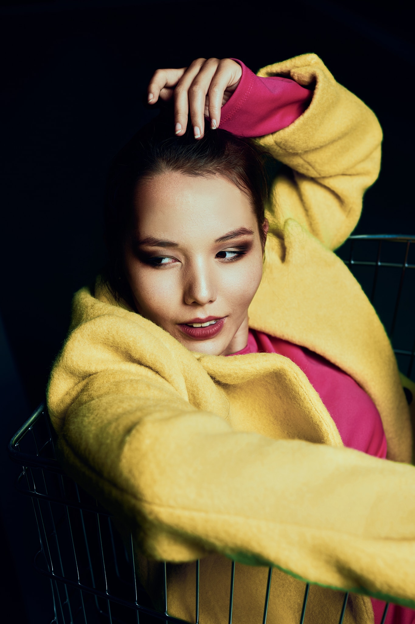 Une femme vêtue d'un manteau jaune vif et d'un haut rose est allongée dans un caddie, regardant sur le côté avec une expression détendue et confiante. Une main est levée sur sa tête et l'éclairage crée des ombres dramatiques.