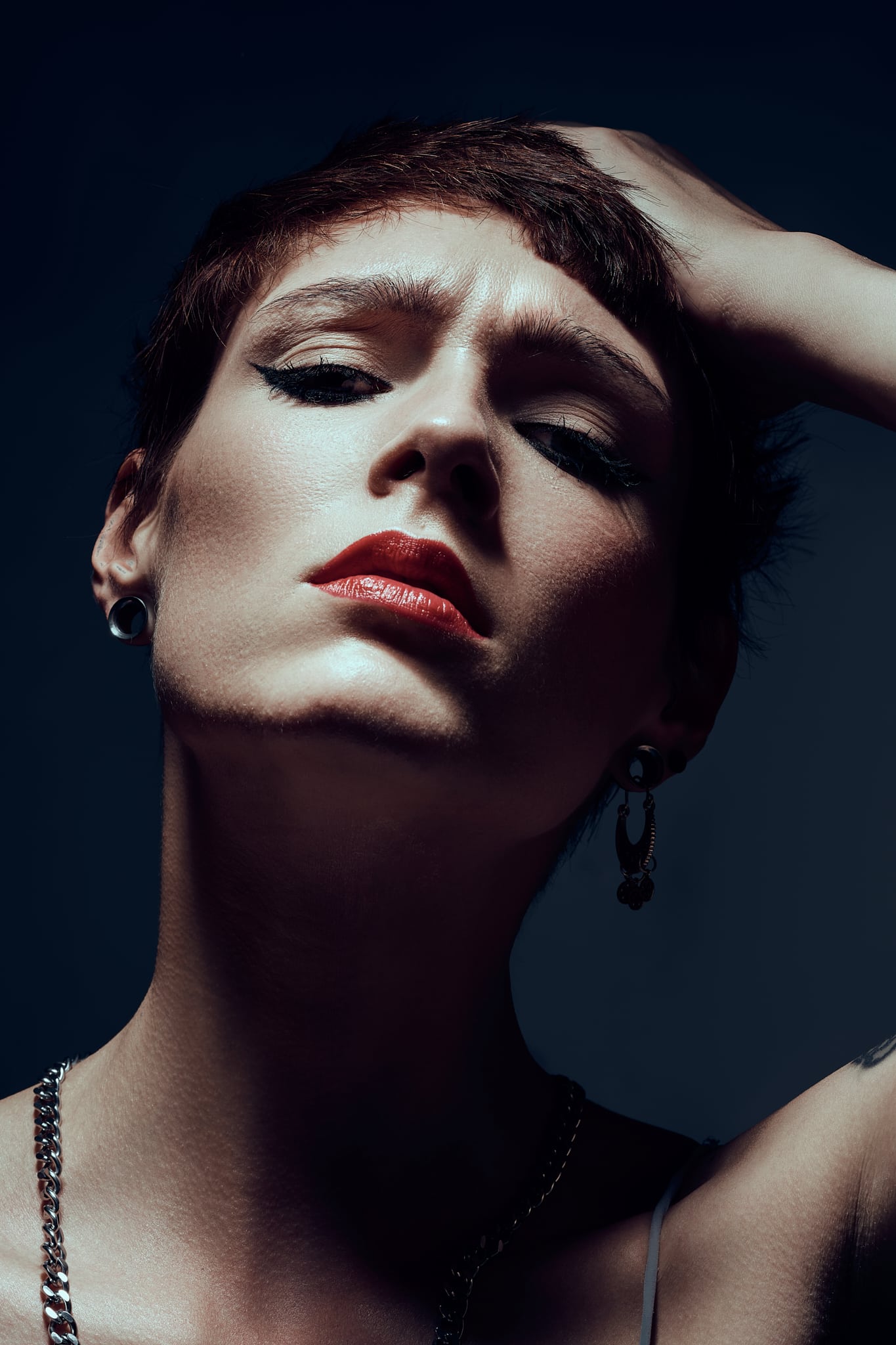 Une personne aux cheveux courts et au maquillage audacieux regarde directement la caméra avec une expression sérieuse, la tête penchée en arrière, une main sur la tête, et un éclairage dramatique projetant des ombres sur le visage et le cou.