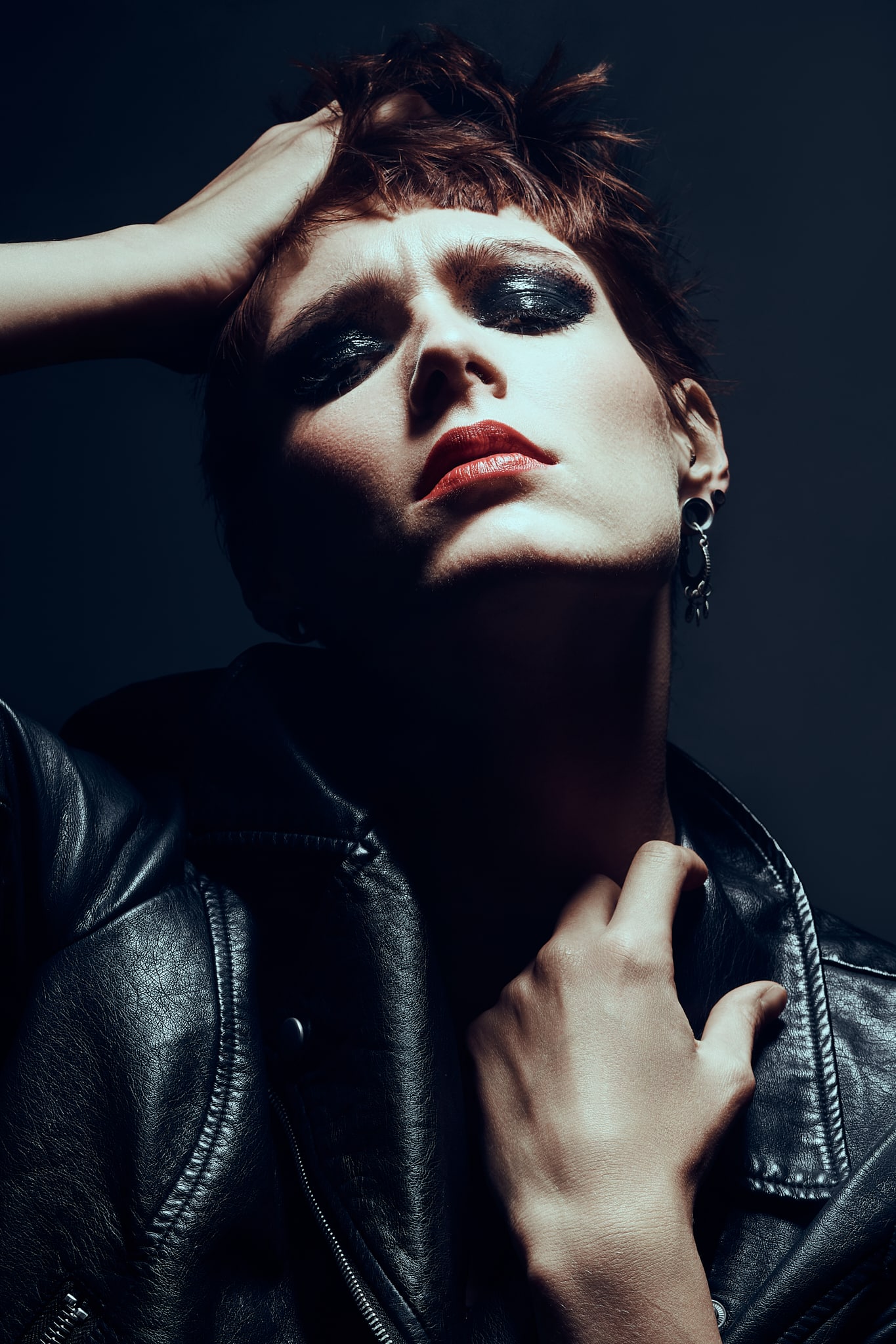 Une personne aux cheveux courts et au maquillage spectaculaire pose dans une veste en cuir noir, une main sur la tête et l'autre sur le cou, regardant vers le haut avec des lèvres rouges et un fard à paupières foncé sur un fond sombre.