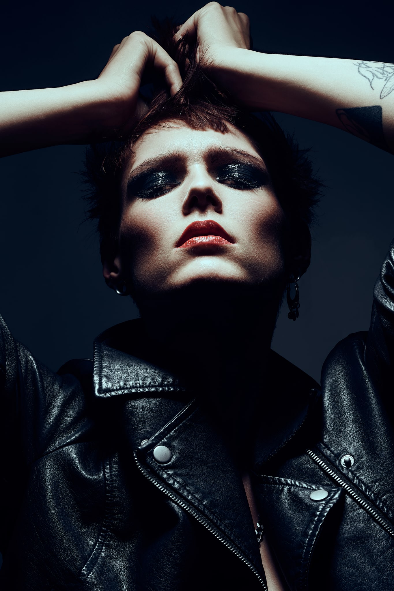 Une personne aux yeux maquillés et au rouge à lèvres rouge prend une pose théâtrale, regardant vers le haut, les mains dans les cheveux courts. Elle porte une veste en cuir noir et arbore un tatouage sur le bras. L'éclairage projette des ombres profondes, créant une atmosphère morose.