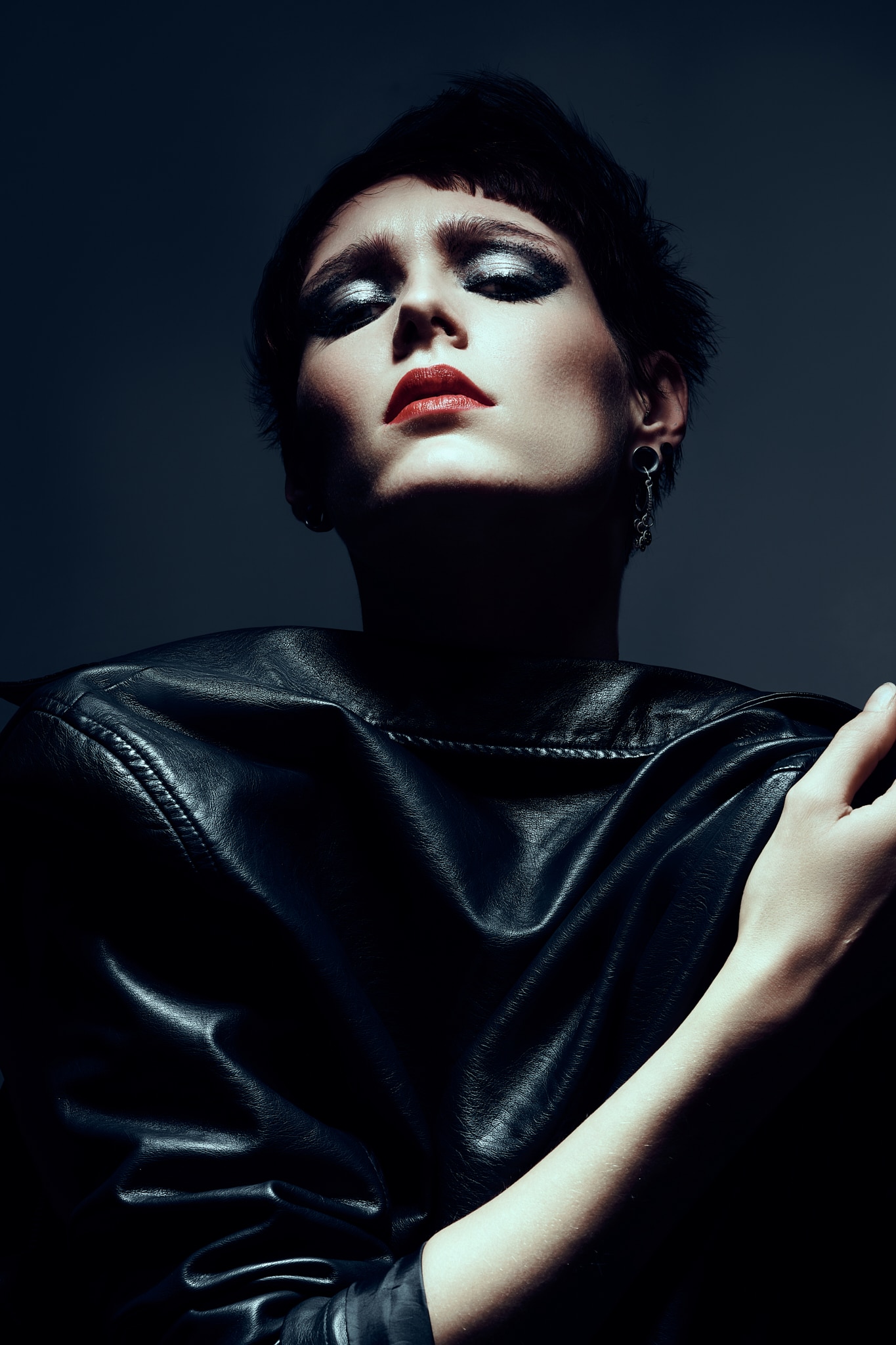 Beauté. Une personne aux cheveux noirs courts et au maquillage spectaculaire pose dans un blouson de cuir noir, les yeux fermés et la tête inclinée vers l'arrière, sous un éclairage sombre qui projette des ombres profondes sur son visage et son corps.