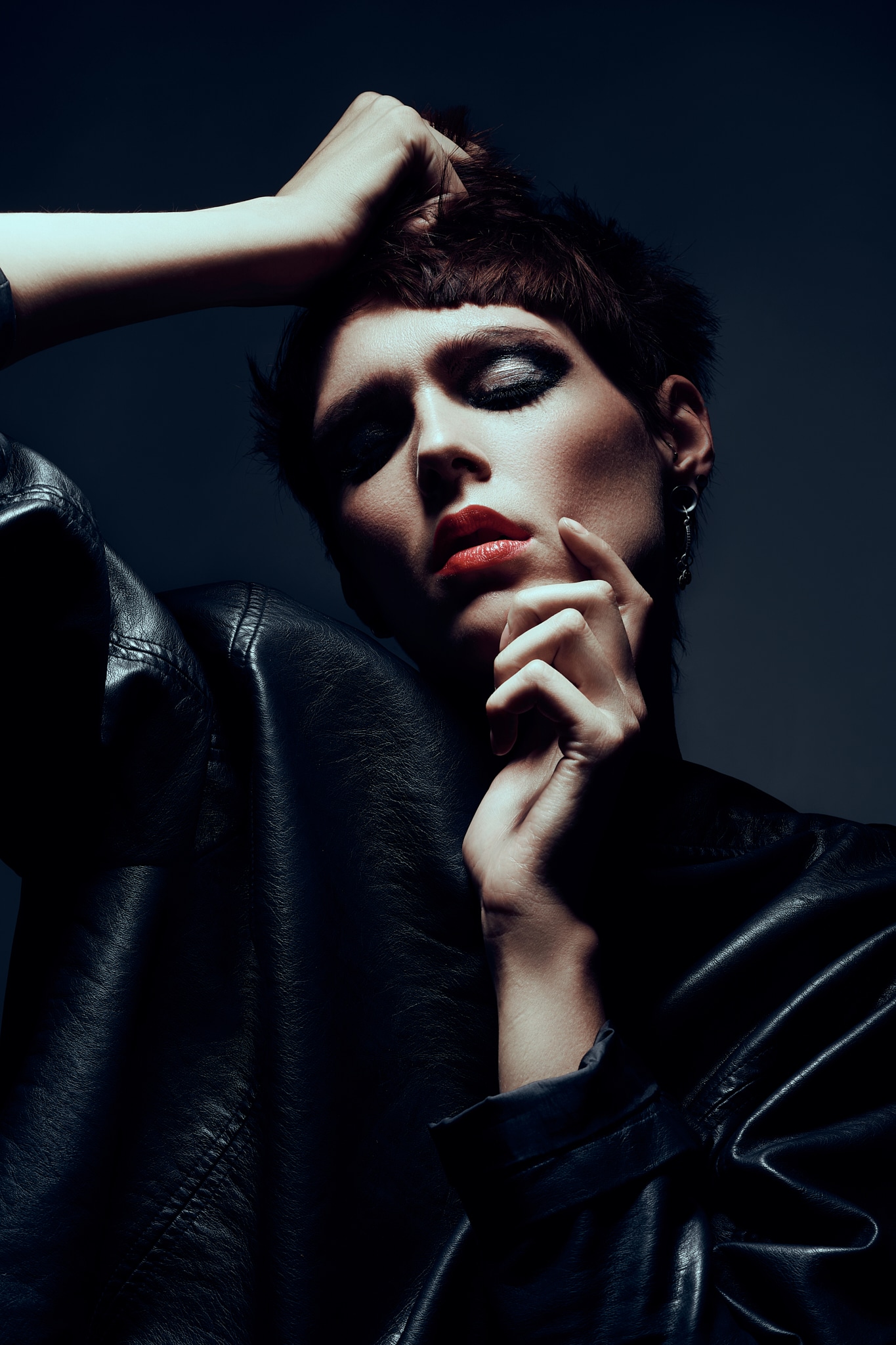 Une personne aux cheveux courts et au maquillage audacieux prend une pose théâtrale dans un blouson de cuir noir, une main sur la tête et l'autre touchant le menton, sur un fond sombre.