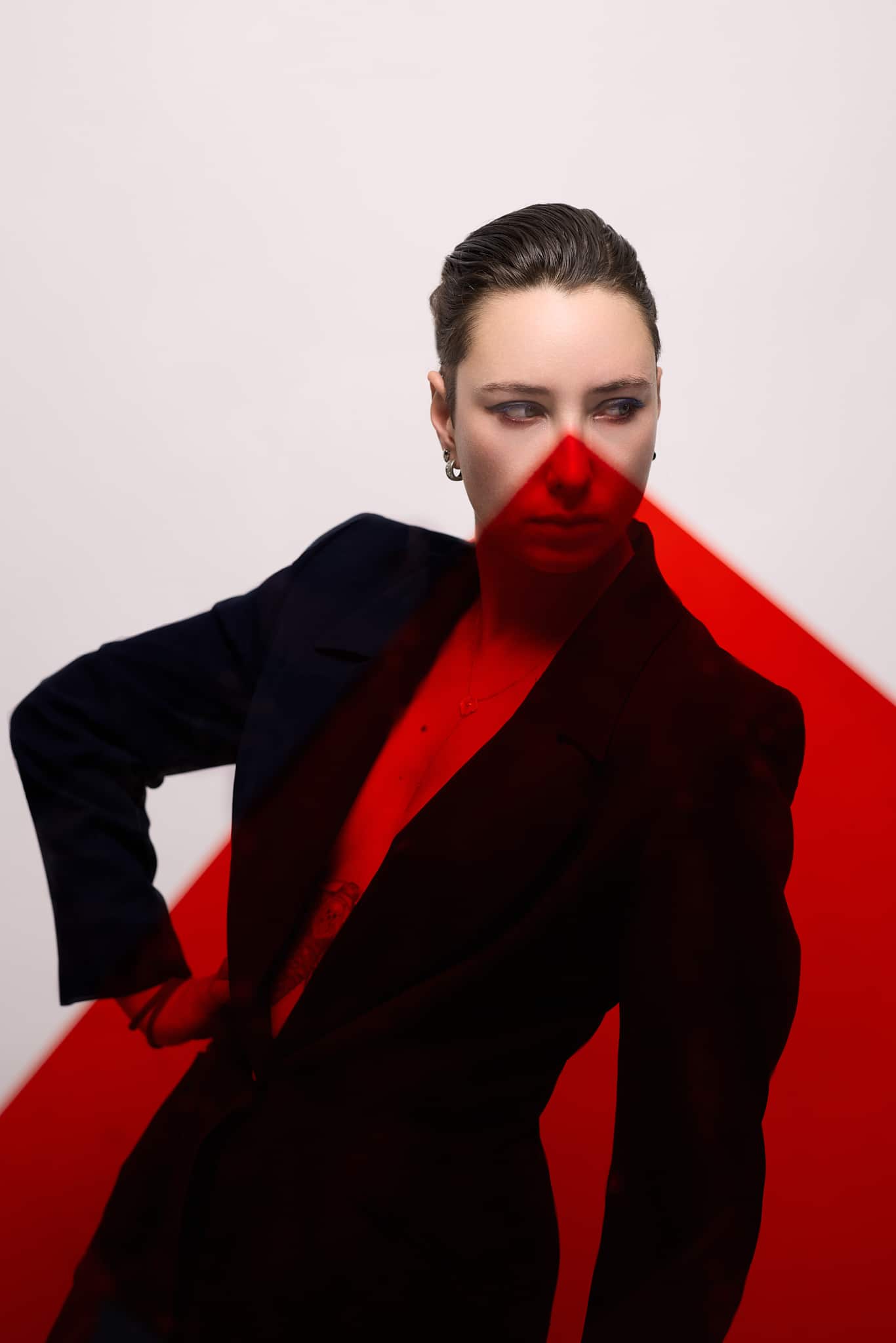 Une personne aux cheveux courts portant un blazer sombre se tient sur un fond blanc, partiellement couvert par une forme géométrique rouge audacieuse, projetant une ombre rouge dramatique sur son visage et son corps.