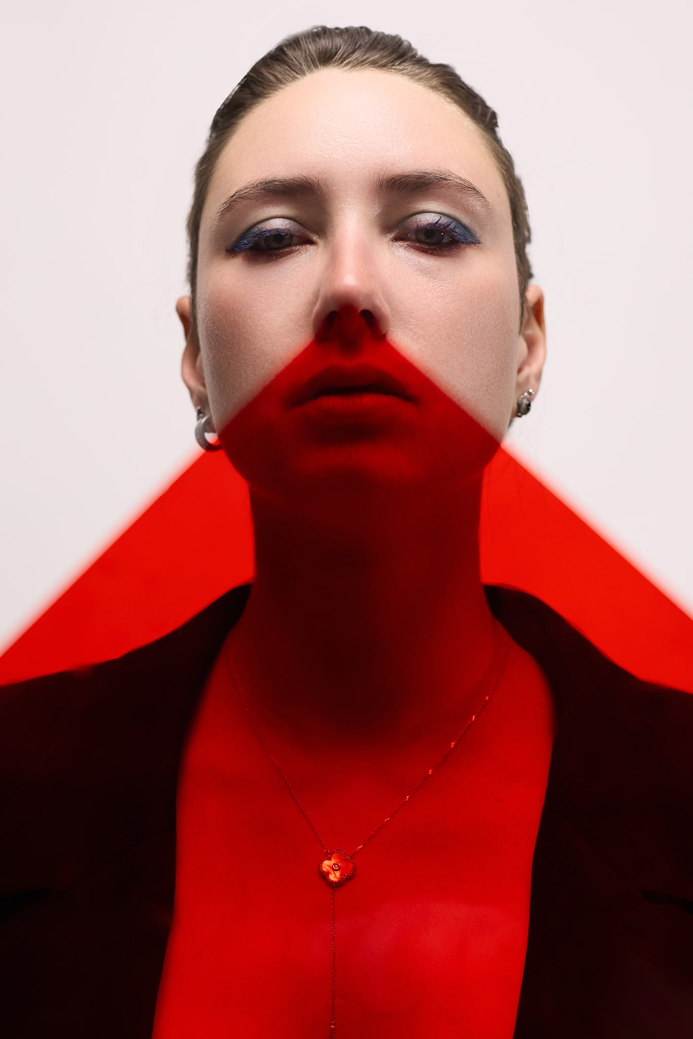 Une femme aux cheveux gominés et au fard à paupières bleu regarde directement la caméra. Un triangle de lumière rouge couvre le bas de son visage et le haut de son corps. Elle porte des boucles d'oreilles, un collier et une veste sombre sur un fond uni.