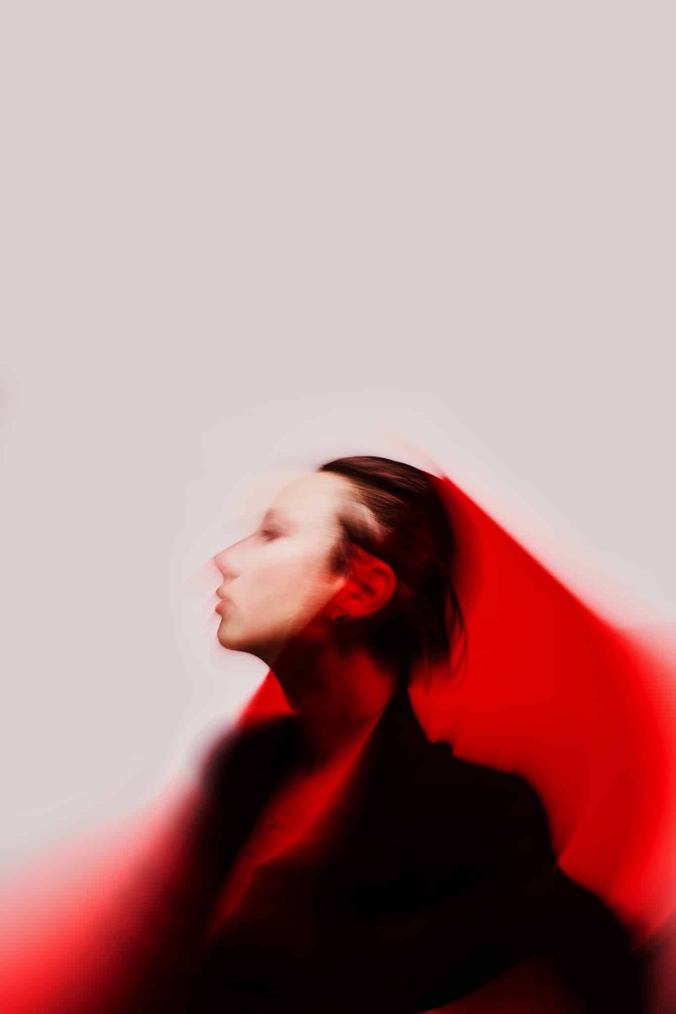 Portrait. Image floue d'une personne aux cheveux longs, tournée vers la gauche, sur un fond uni avec une lumière rouge dramatique qui projette des ombres autour d'elle. La photo a une atmosphère abstraite, rêveuse et morose.