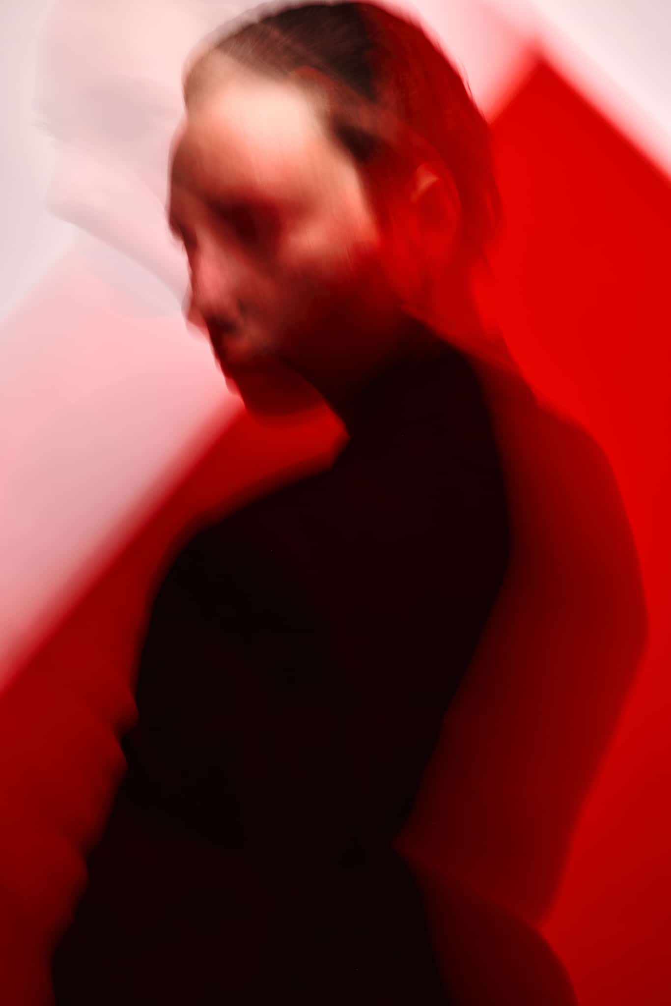 La silhouette floue d'une personne portant des vêtements sombres se détache sur un fond géométrique rouge et blanc, créant un effet abstrait et mystérieux.