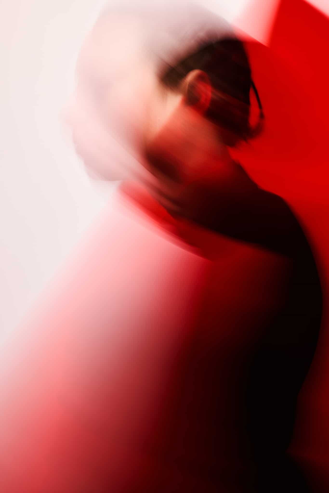 Image abstraite et floue d'une personne aux cheveux noirs de profil, entourée de lumière rouge et blanche, créant une impression de mouvement et une atmosphère onirique.
