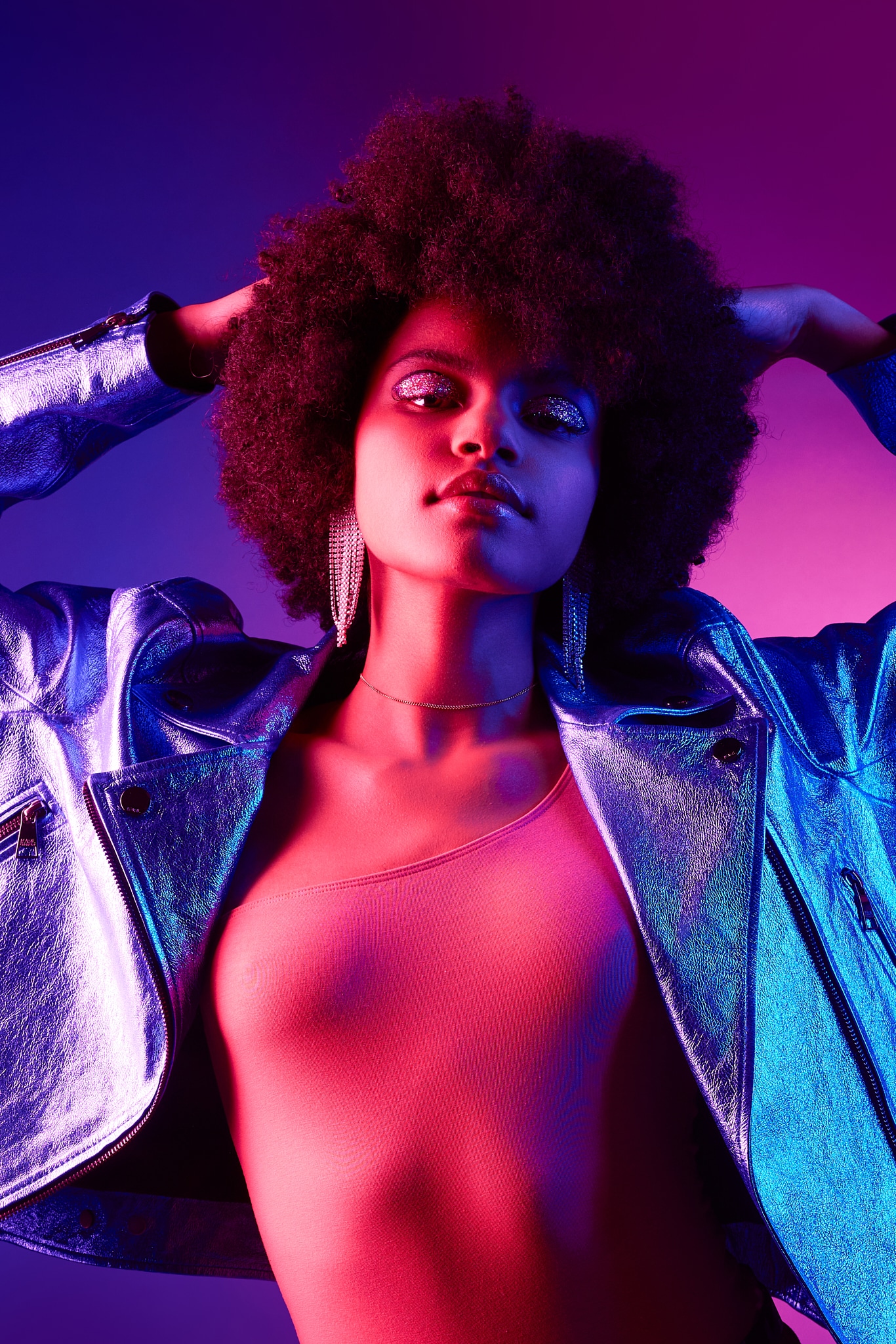 Une femme à la coiffure afro pose avec assurance, les mains derrière la tête. Elle porte une veste métallique brillante, des boucles d'oreilles pendantes et un haut à une épaule, le tout éclairé par une lumière rose et violette vibrante.