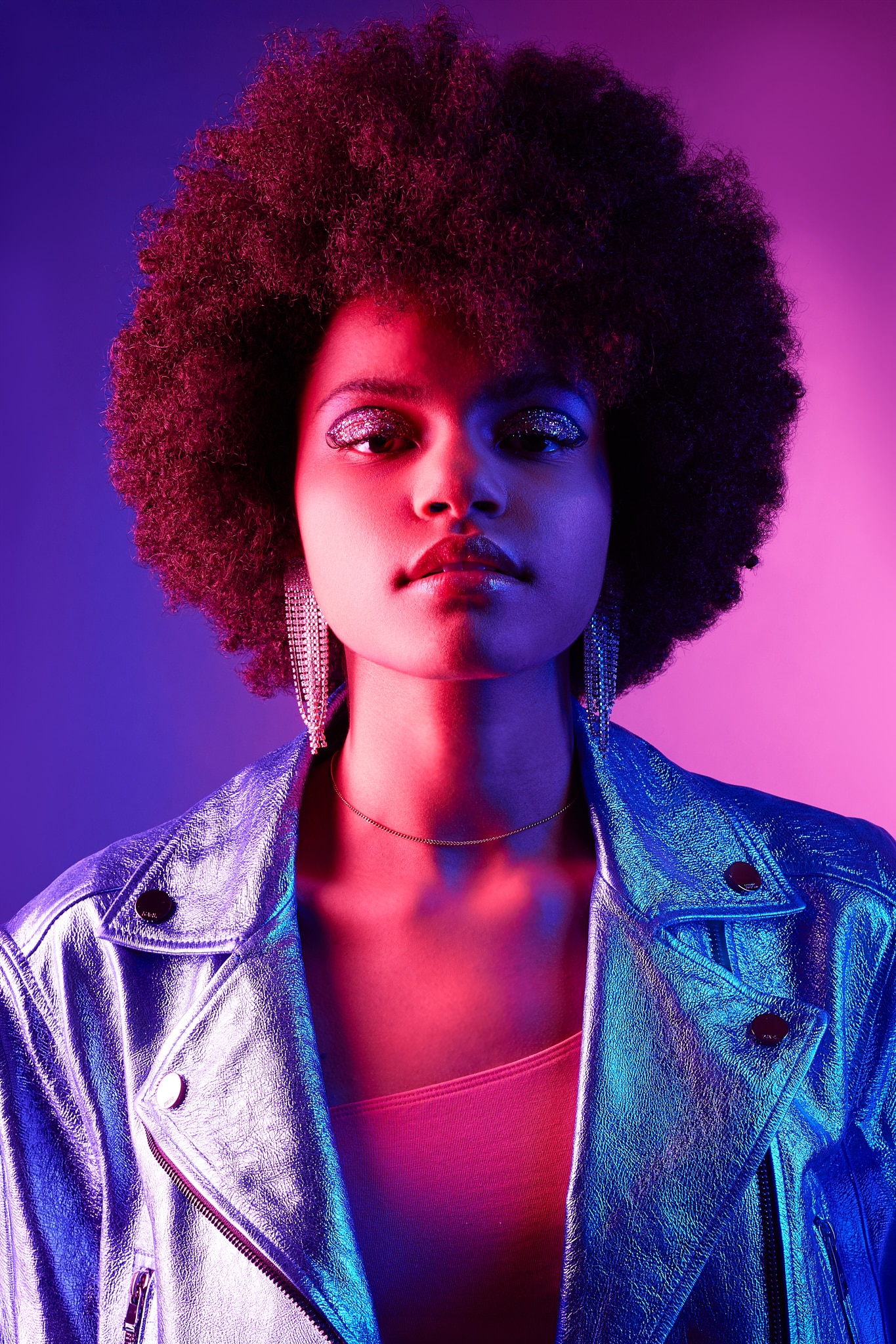 Une femme à la coiffure afro naturelle porte des boucles d'oreilles scintillantes et une veste métallique brillante. Elle se tient debout sur un fond dégradé violet et rose vibrant, éclairé par des lumières colorées.
