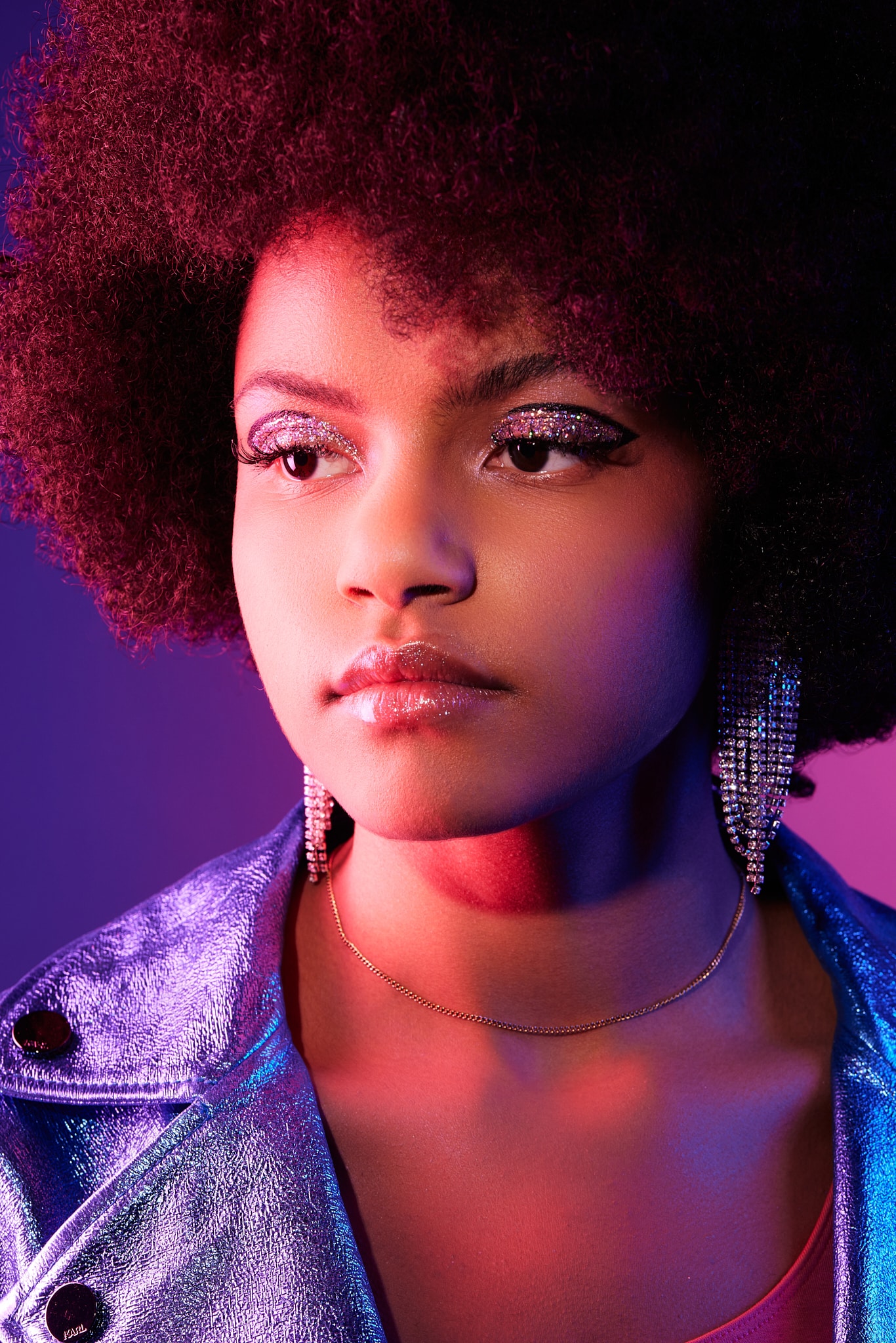Une jeune femme à la coiffure afro naturelle porte un maquillage scintillant pour les yeux, de grandes boucles d'oreilles pendantes, une veste métallique brillante et un collier délicat, le tout éclairé par des lumières roses et bleues vibrantes.