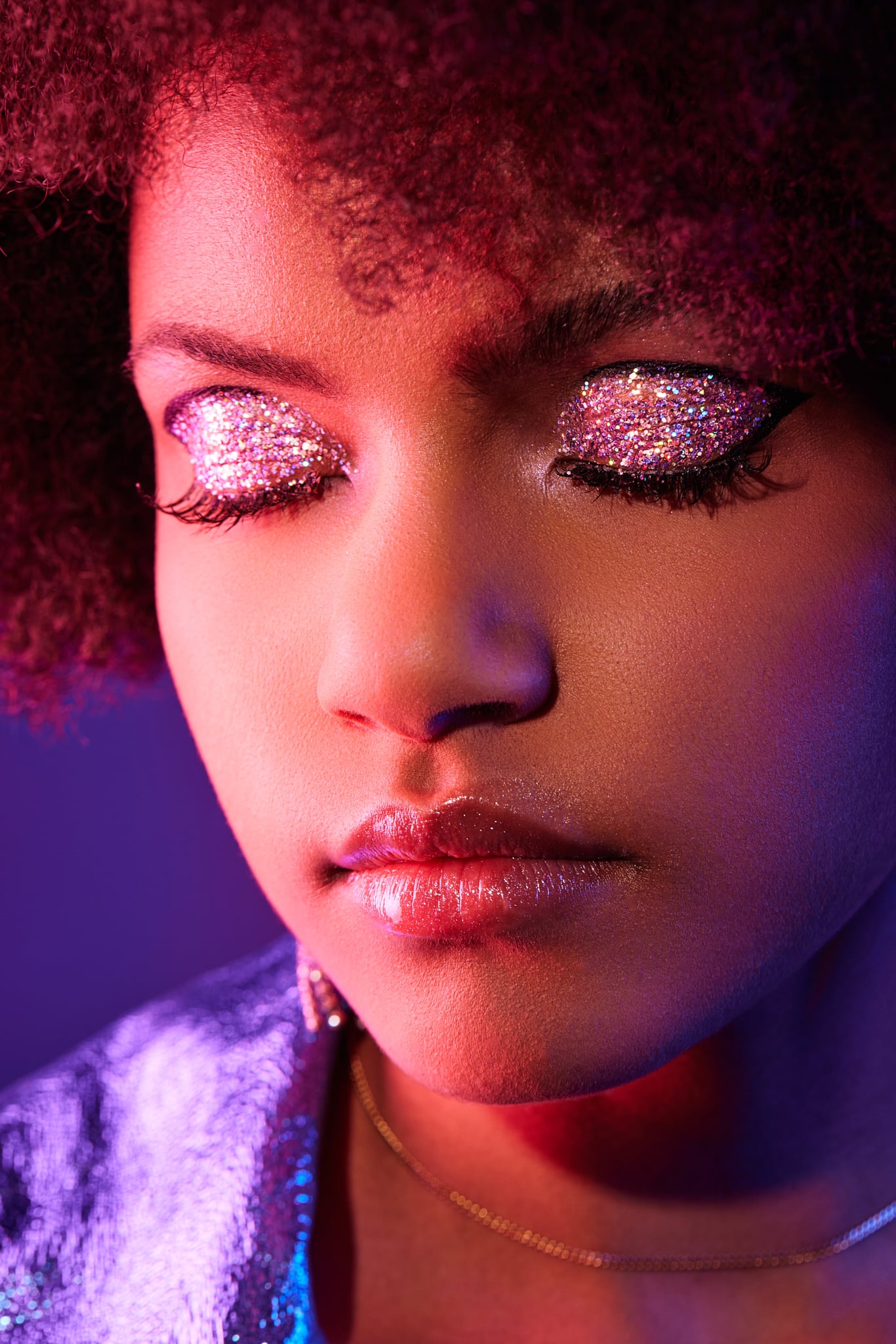 Gros plan d'une personne aux yeux fermés, maquillée de rose pailleté et vêtue d'une tenue violette scintillante, baignant dans un éclairage coloré.