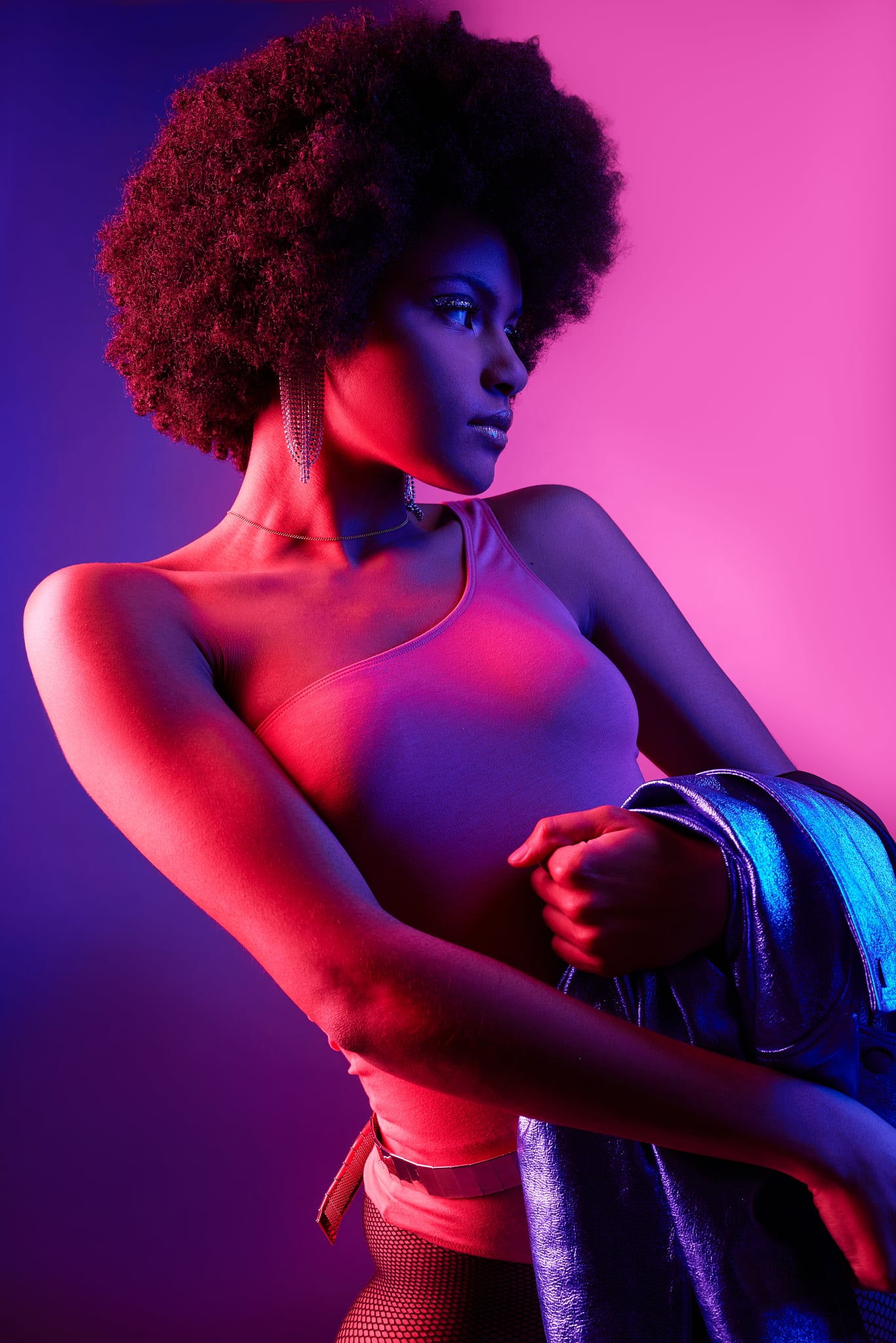 Une femme à la coiffure afro pose avec assurance sous un éclairage dramatique rose et violet, portant un haut à une épaule et une veste brillante au bras, évoquant un style audacieux d'inspiration rétro. Dancing.