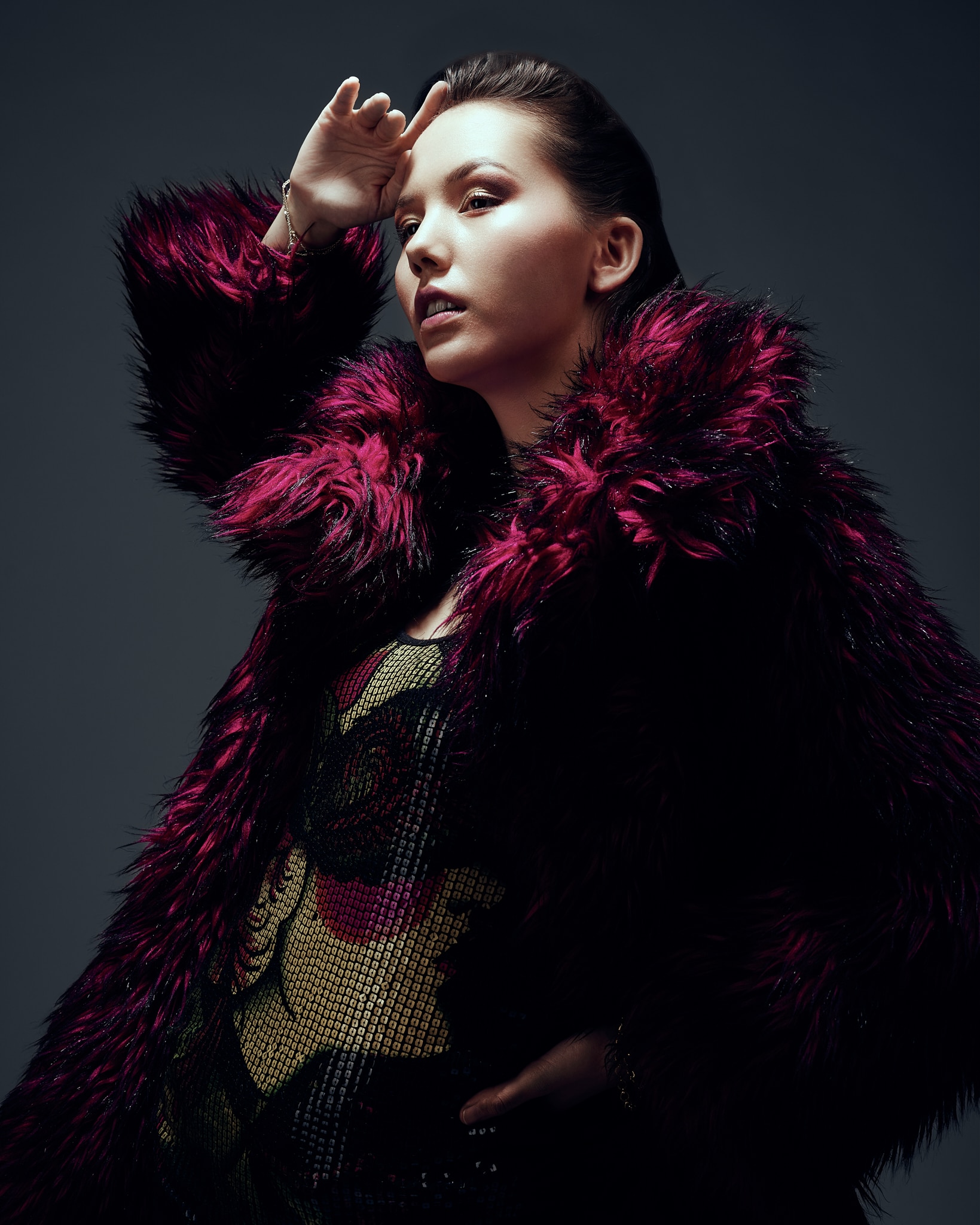 Une femme vêtue d'un luxueux manteau de fausse fourrure bordeaux profond pose, une main sur la hanche et l'autre portée au front, le regard tourné vers le haut, sur un fond sombre et morose.