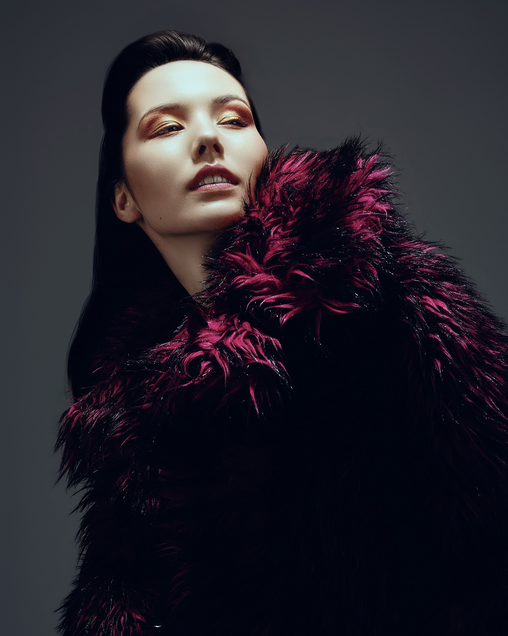 Une personne aux longs cheveux noirs pose dans un décor dramatique et sombre, vêtue d'un luxueux manteau à l'épaisse fourrure magenta et noire, les yeux maquillés d'or scintillant, le regard confiant et tourné vers le haut.