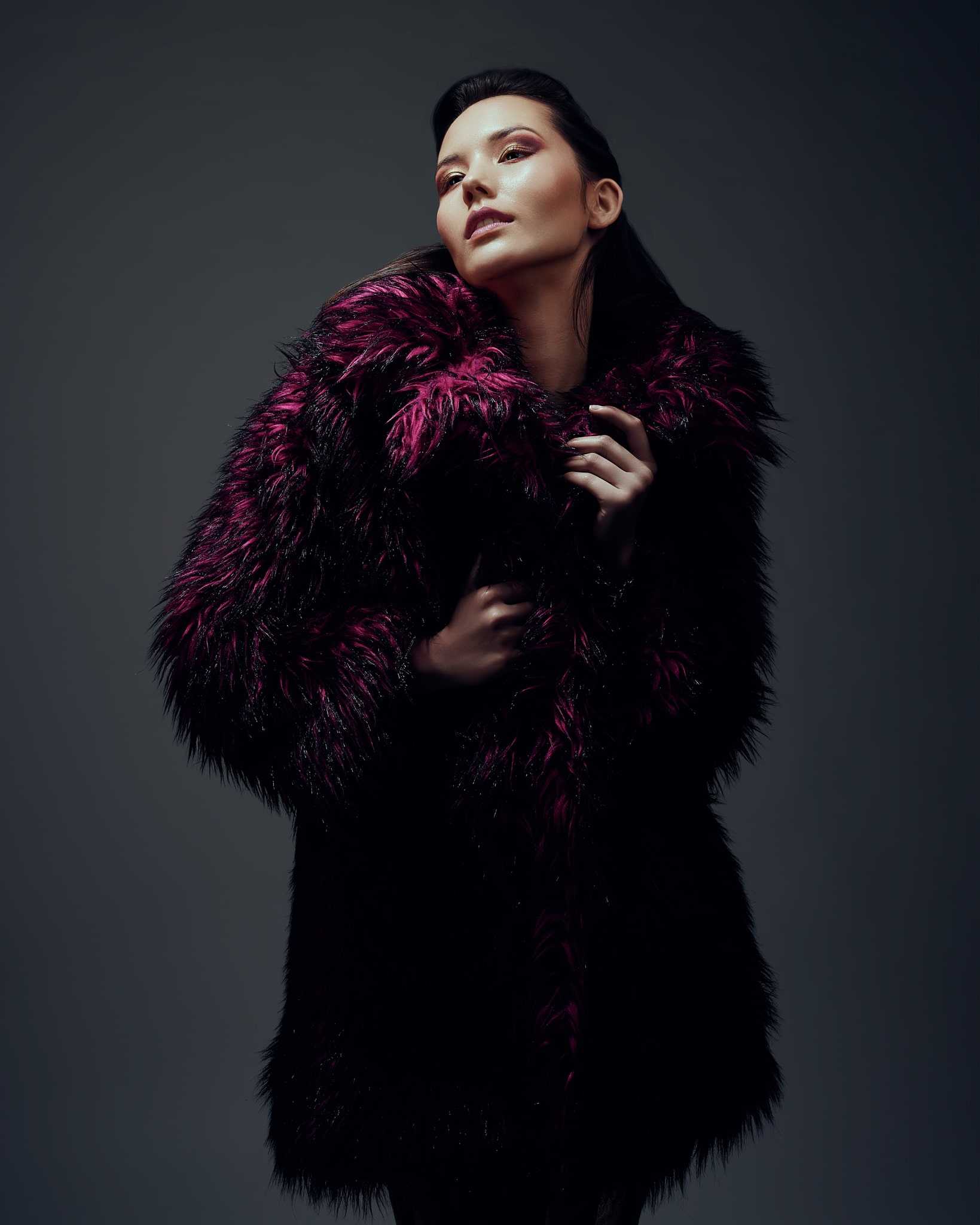 Une femme pose dans un décor sombre et dramatique, vêtue d'un luxueux manteau de fausse fourrure noir et violet foncé. Elle regarde légèrement vers le haut avec une expression confiante, les mains tenant délicatement le col.