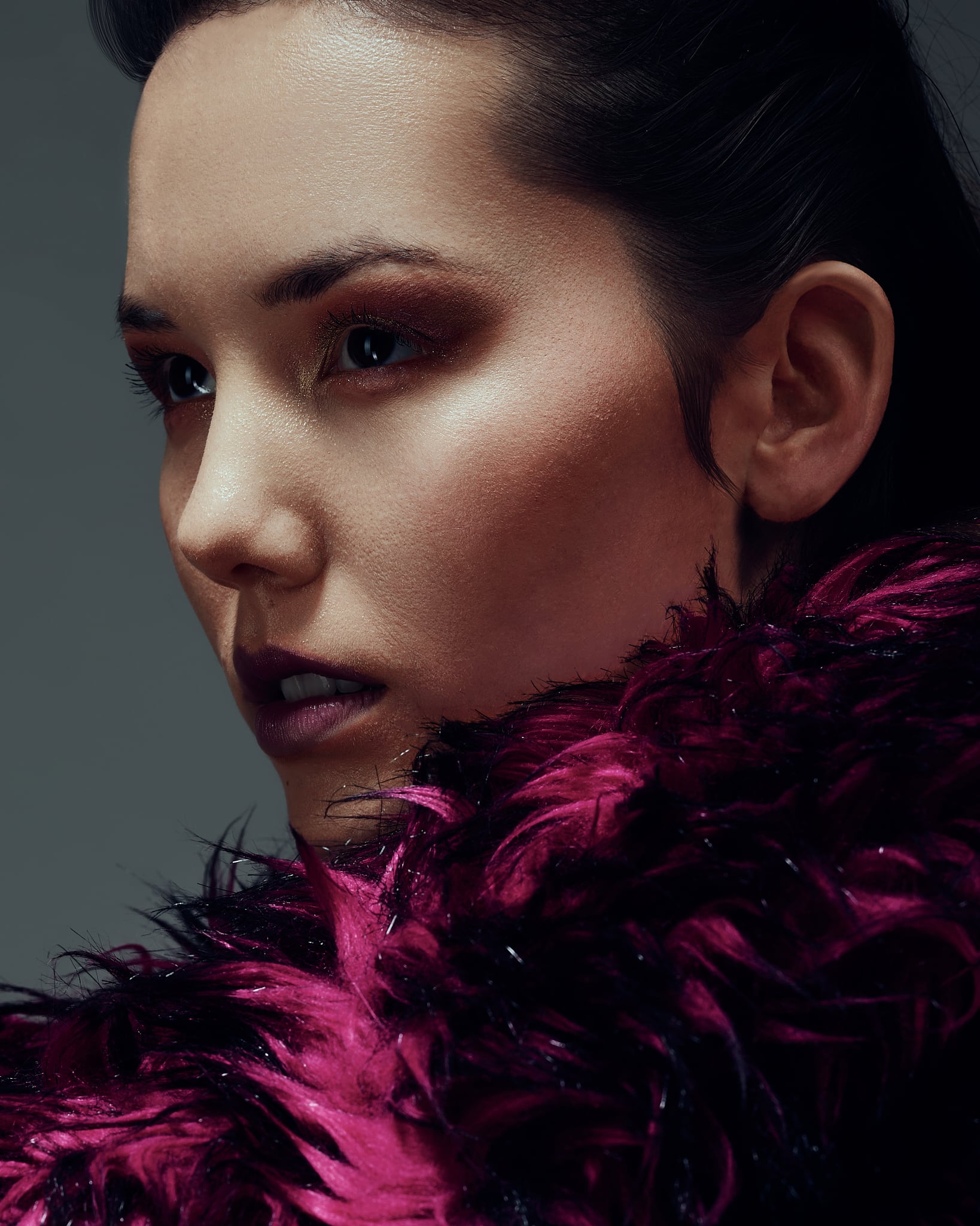 Une femme au maquillage spectaculaire et au rouge à lèvres foncé regarde pensivement sur le côté, son visage partiellement encadré par un luxueux vêtement de plumes d'un magenta profond, sur un fond sombre.