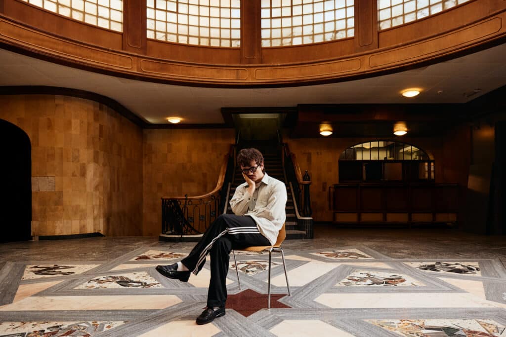 Backstage, A person wearing glasses, a light jacket, and sweatpants sits on a chair in the center of a grand entrance hall featuring marble flooring, a curved staircase, and a large circular skylight.