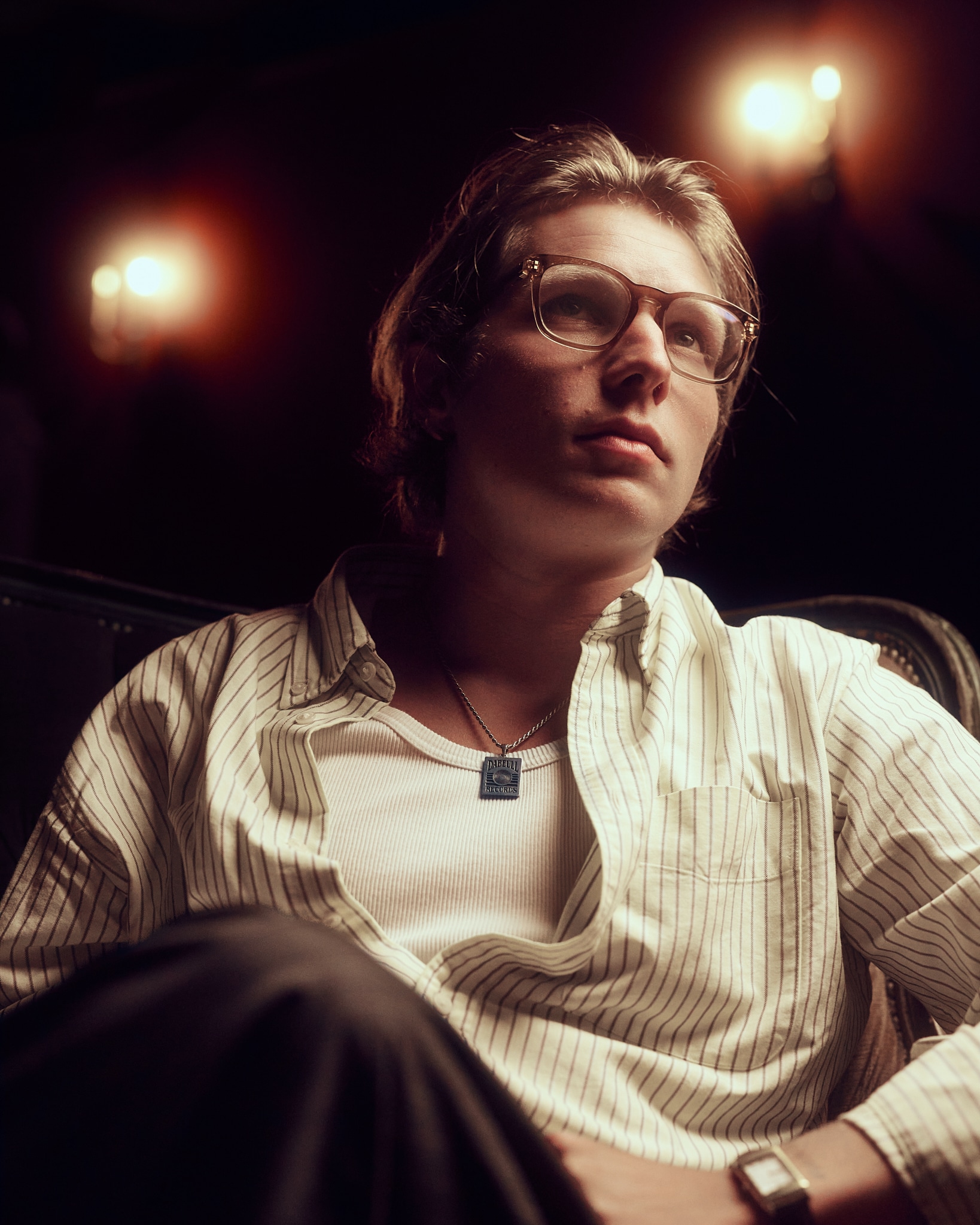 Un jeune homme portant des lunettes, une chemise à rayures sur un maillot de corps blanc et un collier est assis dans une pièce faiblement éclairée par des appliques chaudes en arrière-plan, regardant légèrement vers le haut avec une expression pensive.