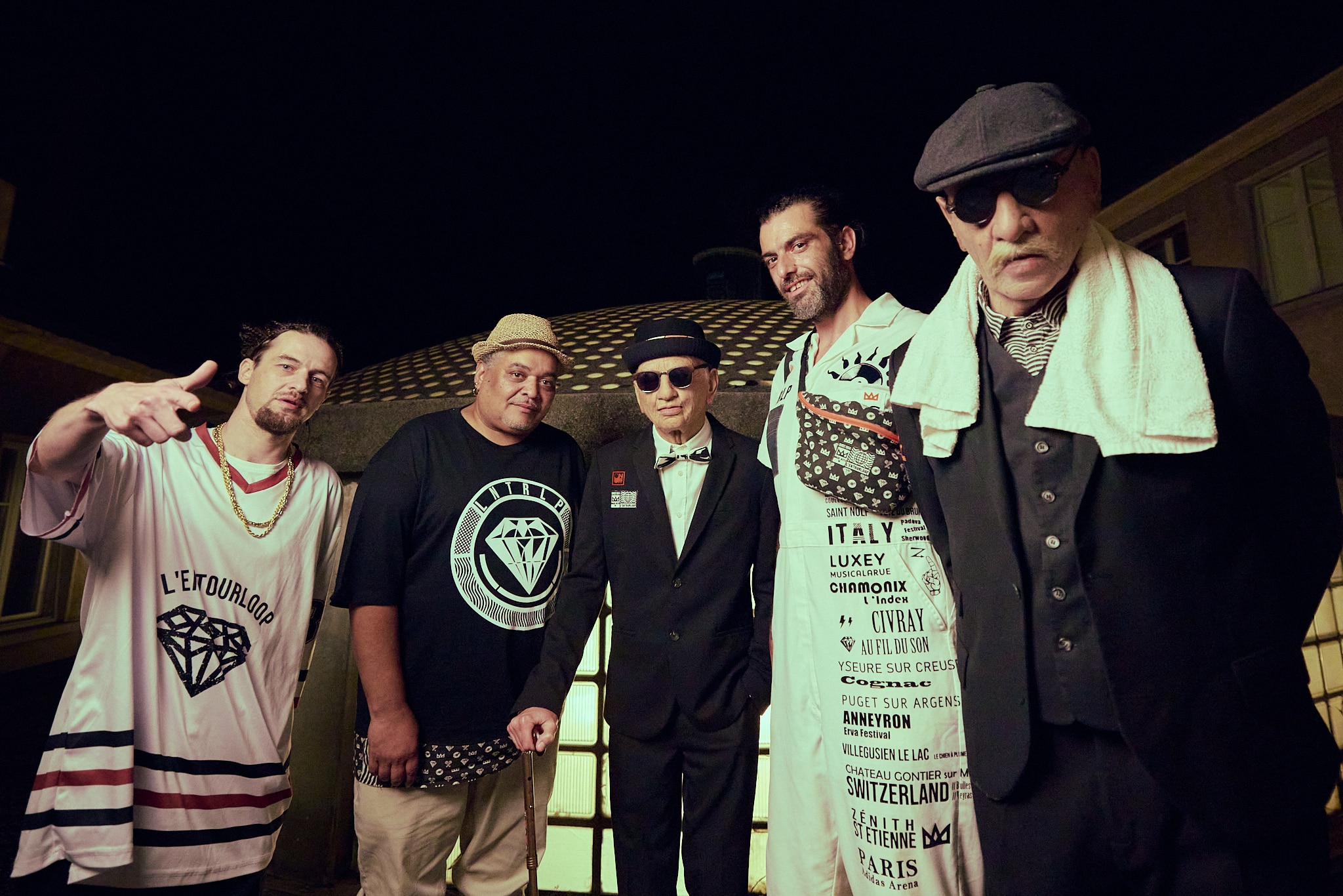 Cinq hommes se tiennent ensemble dans la nuit, habillés dans des styles variés allant du streetwear au vintage. Faisant partie des Gueules de Scène de Francomanias, ils posent avec assurance, certains avec des chapeaux ou des lunettes de soleil, l'un d'entre eux avec une serviette sur l'épaule.