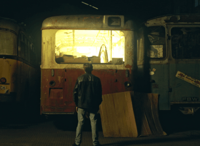 Une personne vêtue d'une veste en cuir se tient à l'écart, face à trois vieux bus rouillés dans un garage faiblement éclairé. Le bus du milieu est éclairé de l'intérieur, jetant une lueur jaune alors que des planches de bois sont appuyées contre lui.
