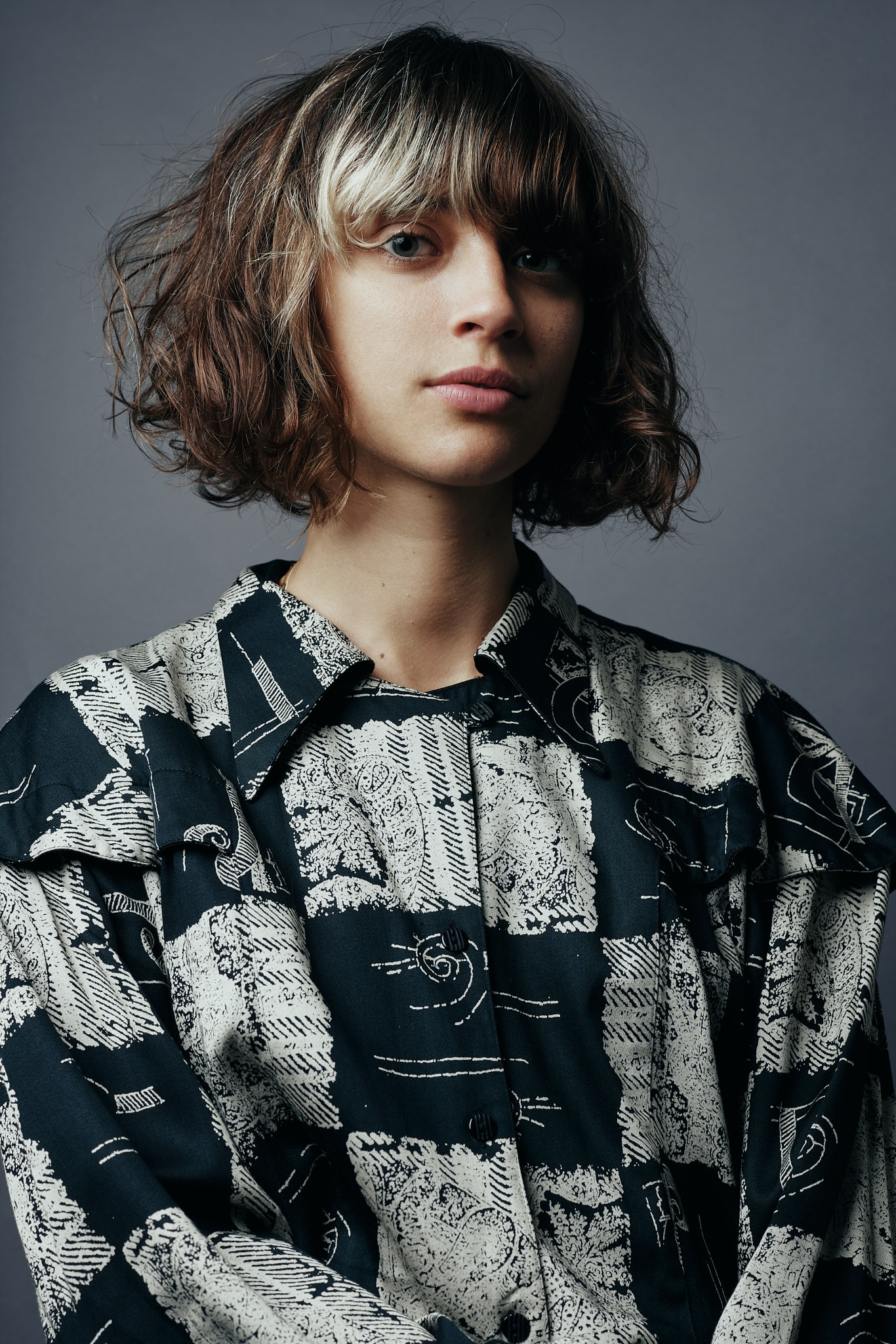 Une personne aux cheveux bruns ondulés, longs comme les épaules, avec une mèche blonde, portant une chemise à motifs noirs et blancs, se tient devant un fond gris uni, illustrant le style unique des Francomanias.