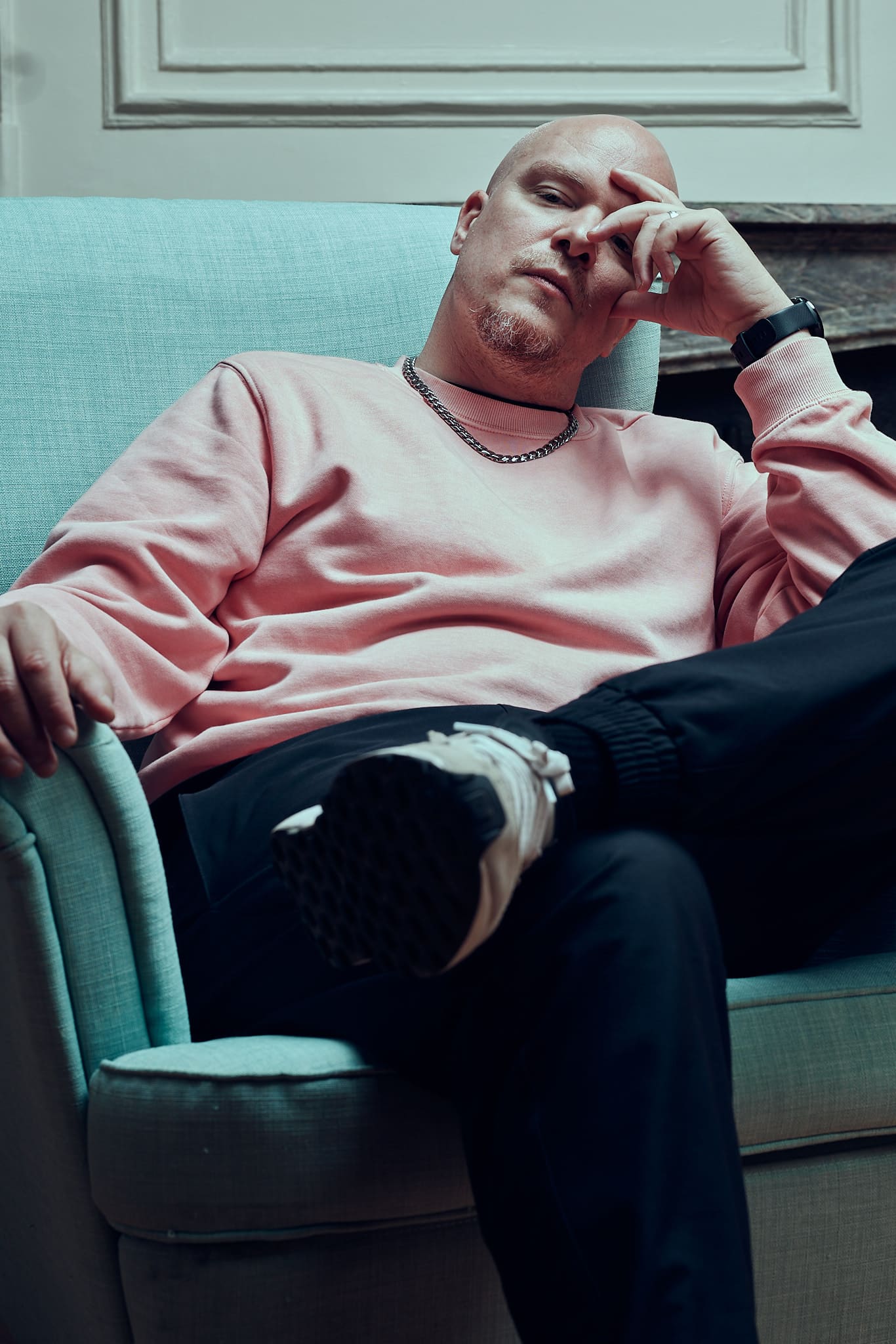 Un homme chauve portant un sweat-shirt rose, un pantalon sombre et des baskets est assis sur un fauteuil bleu clair, une jambe croisée, posant sa tête sur sa main et regardant pensivement la caméra - un vrai moment de Gueules de Scène.