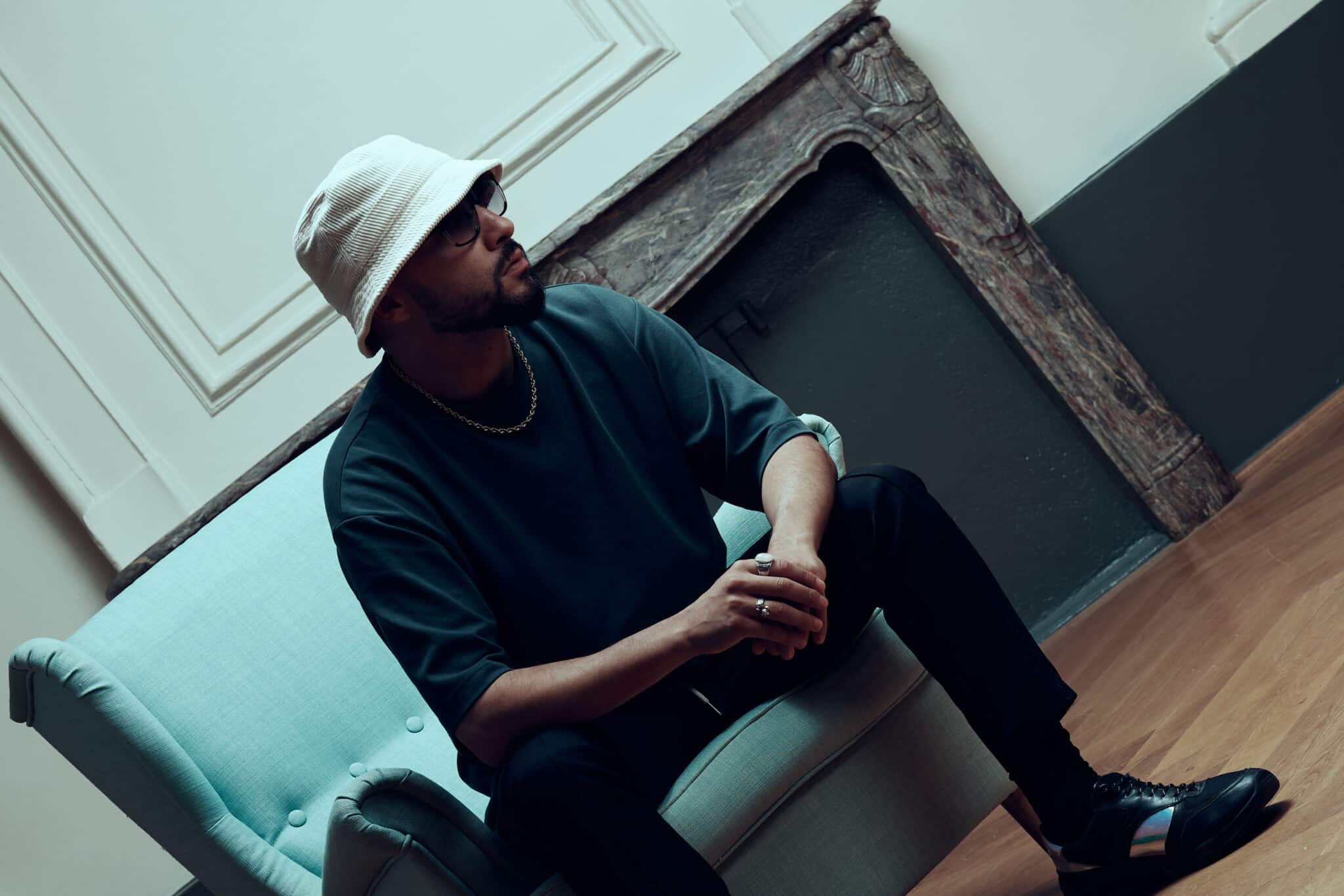 Un homme portant un chapeau blanc, des lunettes et des vêtements sombres est assis sur un canapé bleu clair dans une pièce élégante avec des planchers en bois et des moulures décoratives, incarnant l'ambiance décontractée des Francomanias en regardant sur le côté avec une posture détendue.