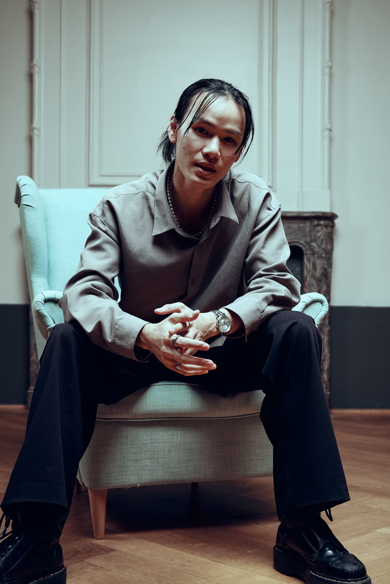 Une personne aux cheveux longs est assise sur un fauteuil de couleur claire, penchée en avant, les mains jointes. Dans ce moment de Gueules de Scène aux Francomanias, ils regardent directement la caméra dans une pièce faiblement éclairée au sol en bois.