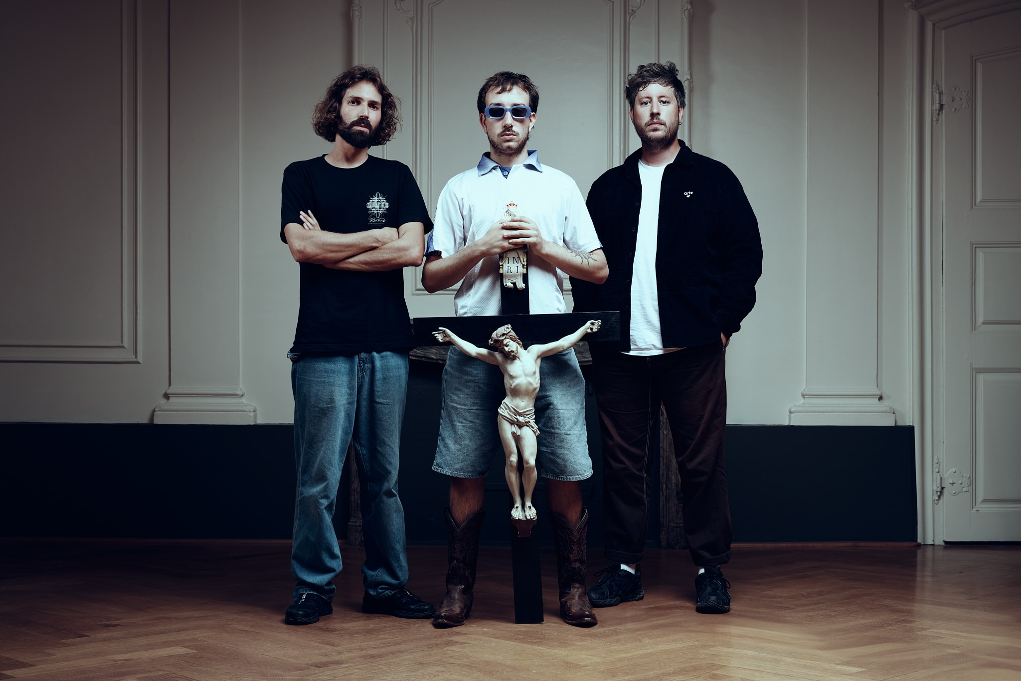 Trois hommes se tiennent à l'intérieur sur un plancher en bois. L'homme du milieu, portant des lunettes de soleil et tenant un crucifix, est flanqué de deux hommes habillés de manière décontractée. Derrière eux, un mur blanc uni et des panneaux de porte évoquent l'esprit des Gueules de Scène aux Francomanias.