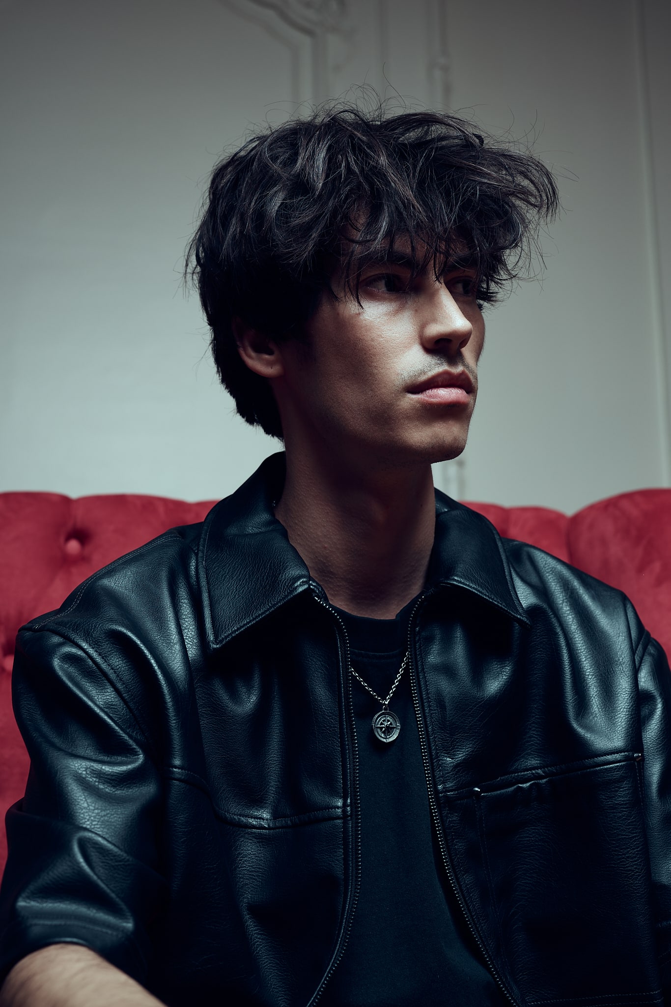 Backstage, Un jeune homme aux cheveux noirs ébouriffés, portant une veste en cuir noir et un collier, est assis sur un canapé rouge, regardant pensivement sur le côté dans une pièce doucement éclairée, capturant l'esprit créatif des Gueules de Scène aux Francomanias.