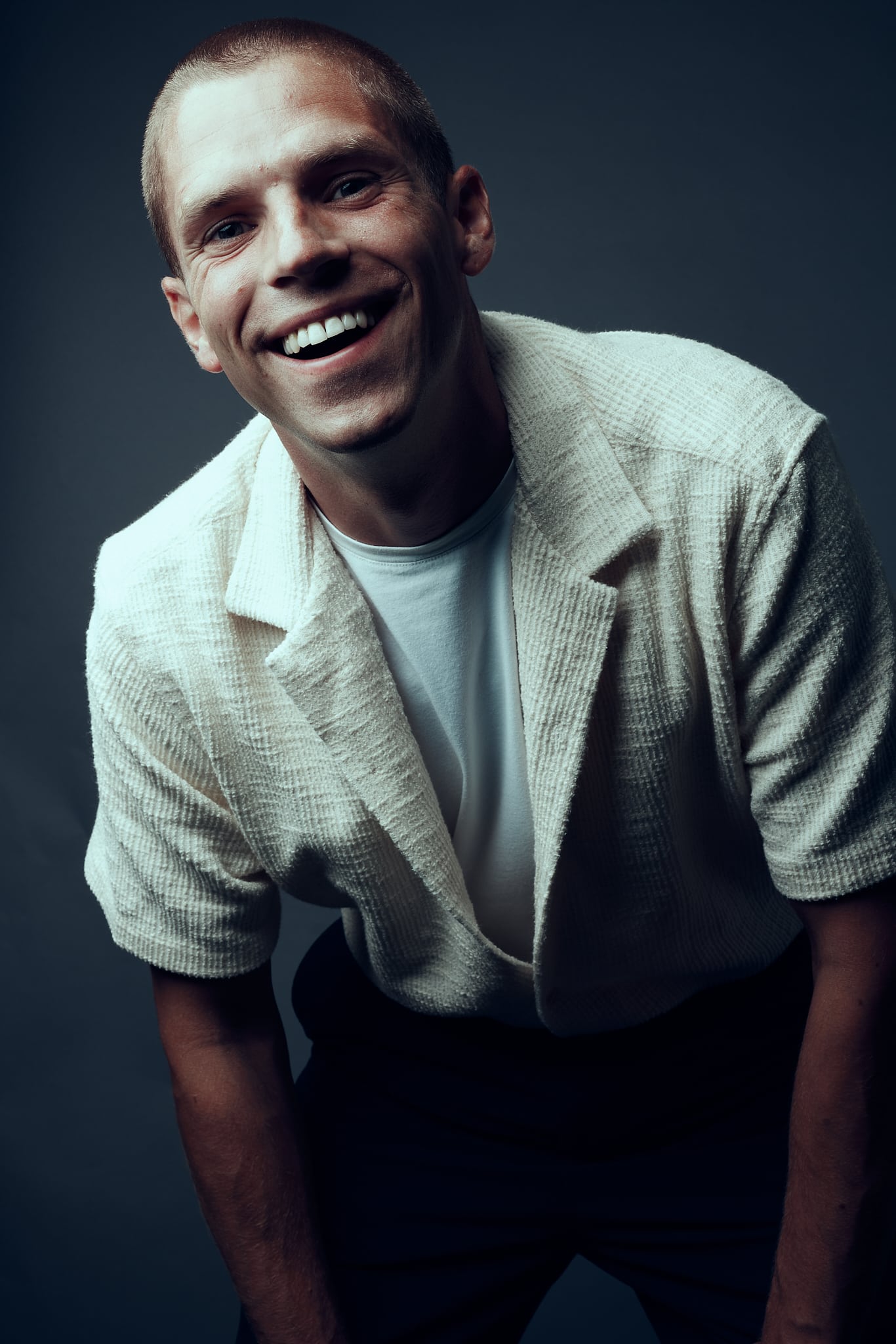 Un homme souriant aux cheveux courts se penche en avant, vêtu d'une veste texturée à manches courtes de couleur crème sur une chemise blanche, incarnant l'esprit des Francomanias, sur un fond gris foncé.