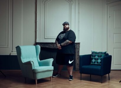 Un homme portant une casquette, un t-shirt et un short noirs se tient à l'écart entre un fauteuil bleu clair et une chaise bleu foncé avec un coussin à motifs dans une pièce élégante aux murs clairs et au sol en bois.