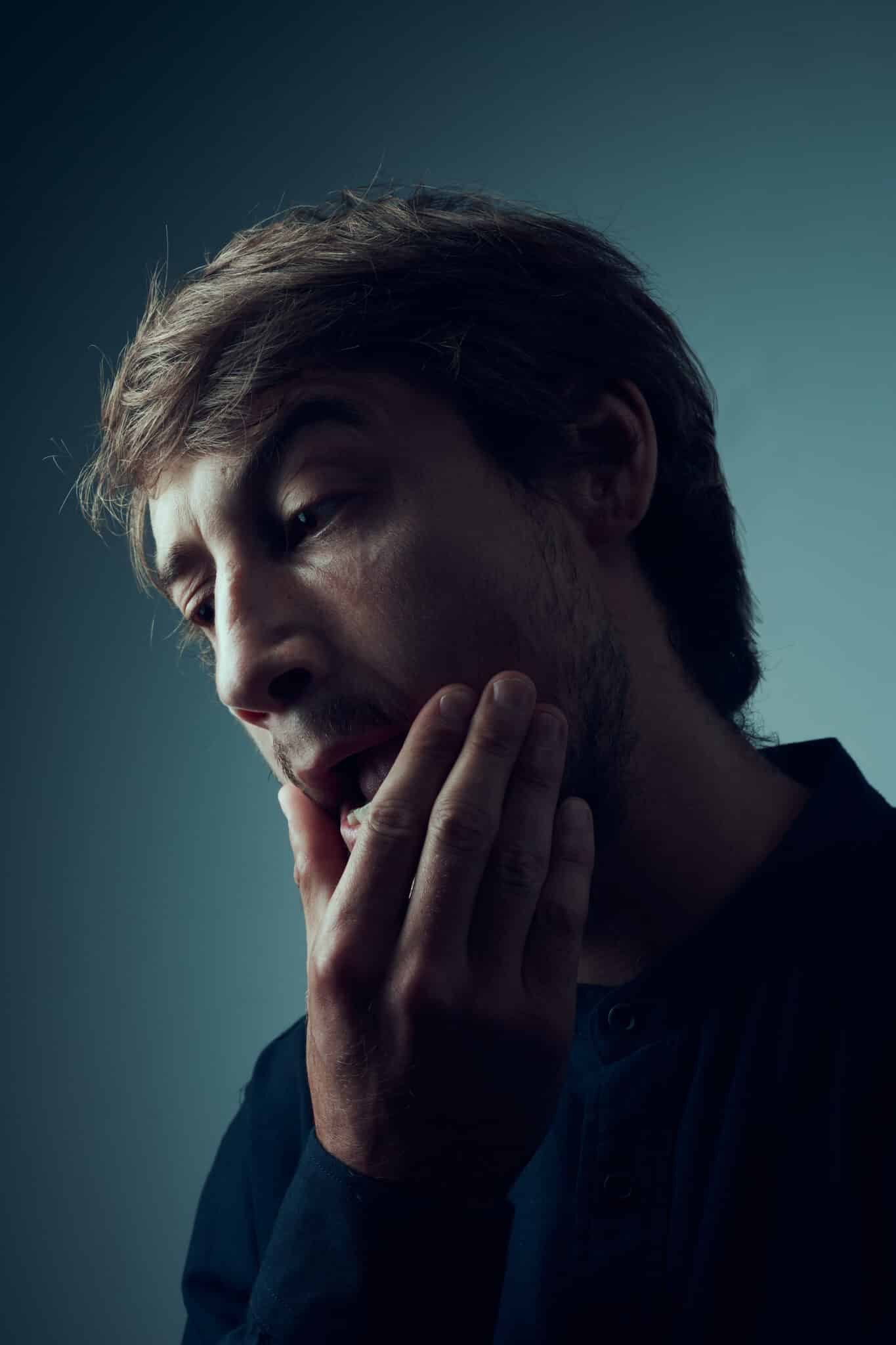 Un homme aux cheveux courts, ressemblant au comédien Luca Colucci, se touche la joue et la mâchoire avec une expression douloureuse, suggérant une gêne dentaire ou maxillaire. Il porte une chemise sombre sur un fond neutre et dégradé.