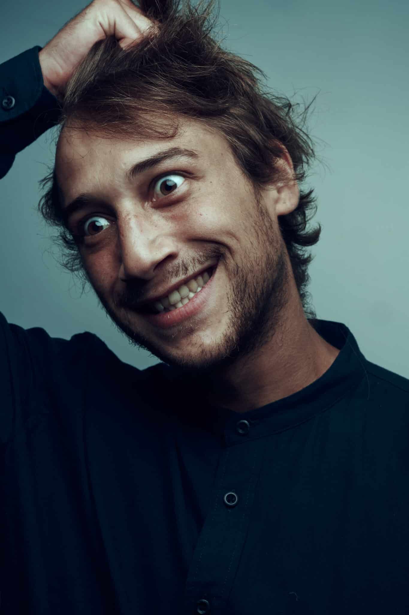 Un homme aux cheveux en désordre affiche un large sourire, les yeux grands ouverts, et se gratte la tête d'une main. Il porte une chemise sombre boutonnée et est éclairé par une lumière douce et lunatique - capturant le charme excentrique d'un comédien comme Luca Colucci sur un fond uni.