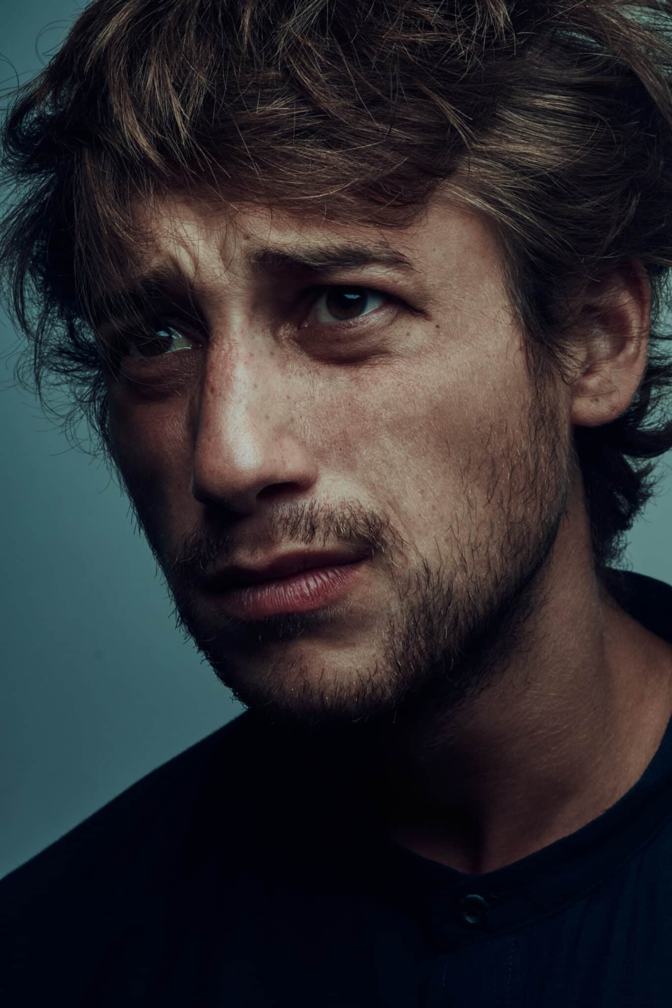 A man with tousled brown hair and light stubble looks upward with a serious and contemplative expression - evoking actor Luca Colucci. He wears a dark shirt against a plain background.