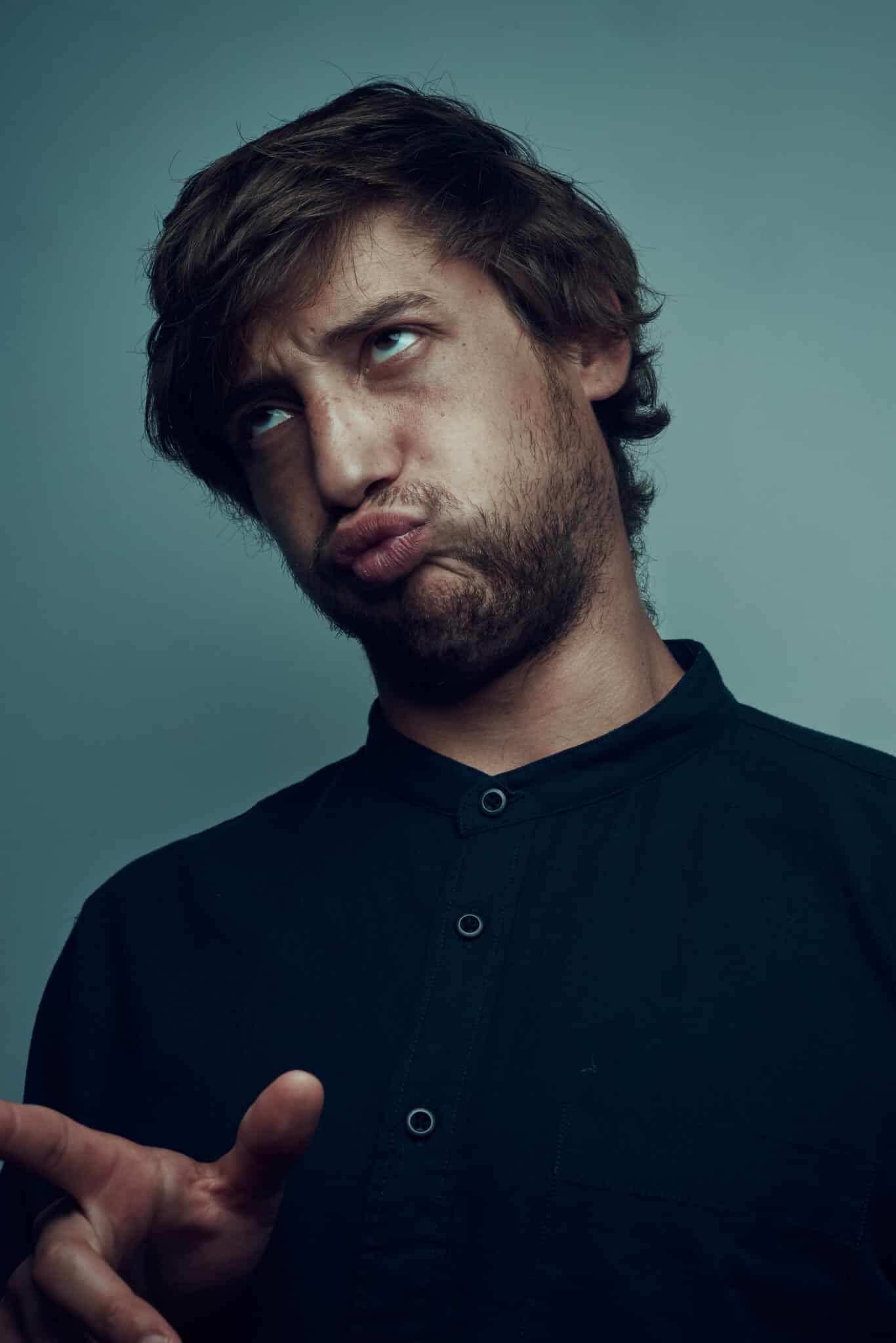 Un homme aux cheveux bruns en désordre et à la barbe fait une grimace ridicule, pinçant les lèvres et haussant les sourcils comme un vrai comédien, tout en pointant du doigt. Il porte une chemise à boutons foncée devant un fond bleu-gris uni.