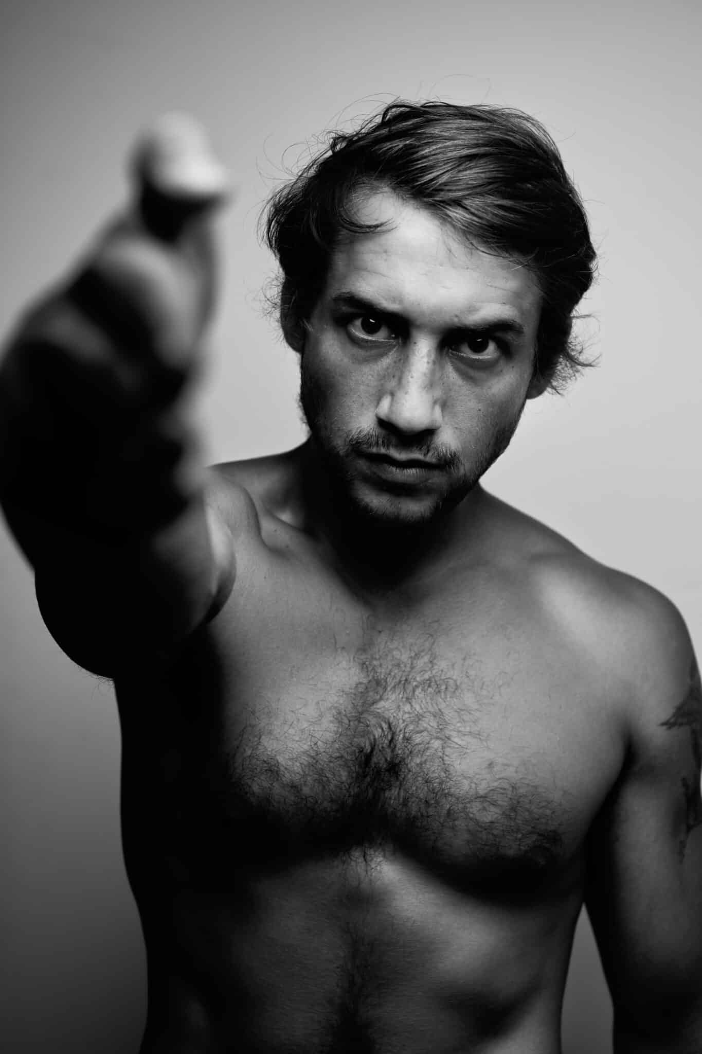 Photo en noir et blanc du comédien Luca Colucci, torse nu, fixant intensément l'appareil photo et tendant le bras vers l'avant avec la main hors champ. Ses cheveux ébouriffés et l'arrière-plan uni ajoutent à l'ambiance dramatique.