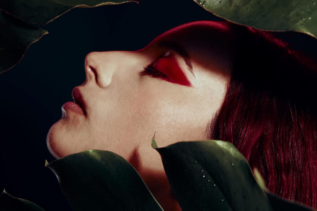 Close-up of a person with red eyes and red hair, surrounded by large green leaves against a dark background.