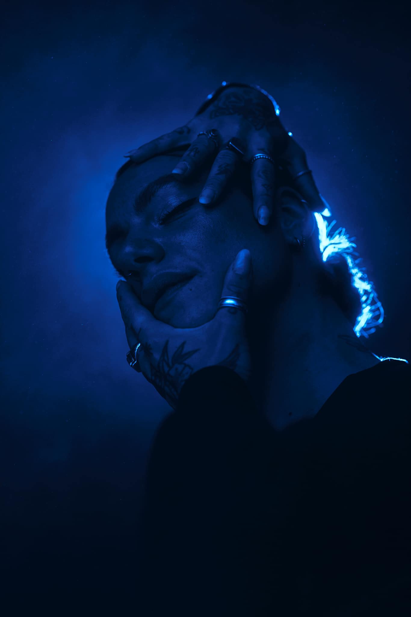 Une personne éclairée par une lumière bleue pose avec ses mains encadrant son visage, mettant en évidence les tatouages et les bagues sur ses doigts. L'arrière-plan est sombre, créant une atmosphère dramatique et morose.