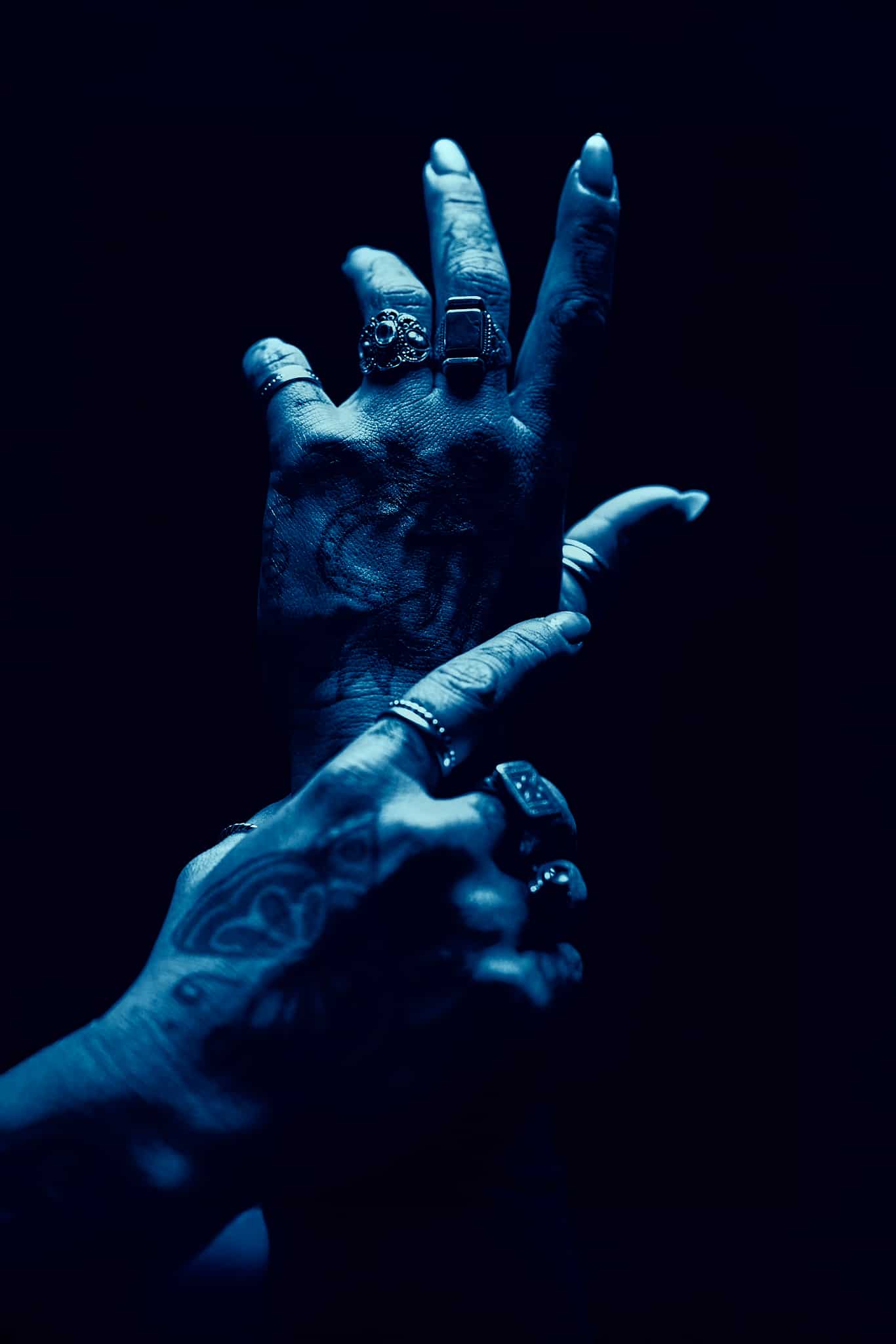 Deux mains tatouées avec de multiples bagues, l'une tenant délicatement le poignet de l'autre, sont éclairées par une lumière bleue dramatique sur un fond sombre.