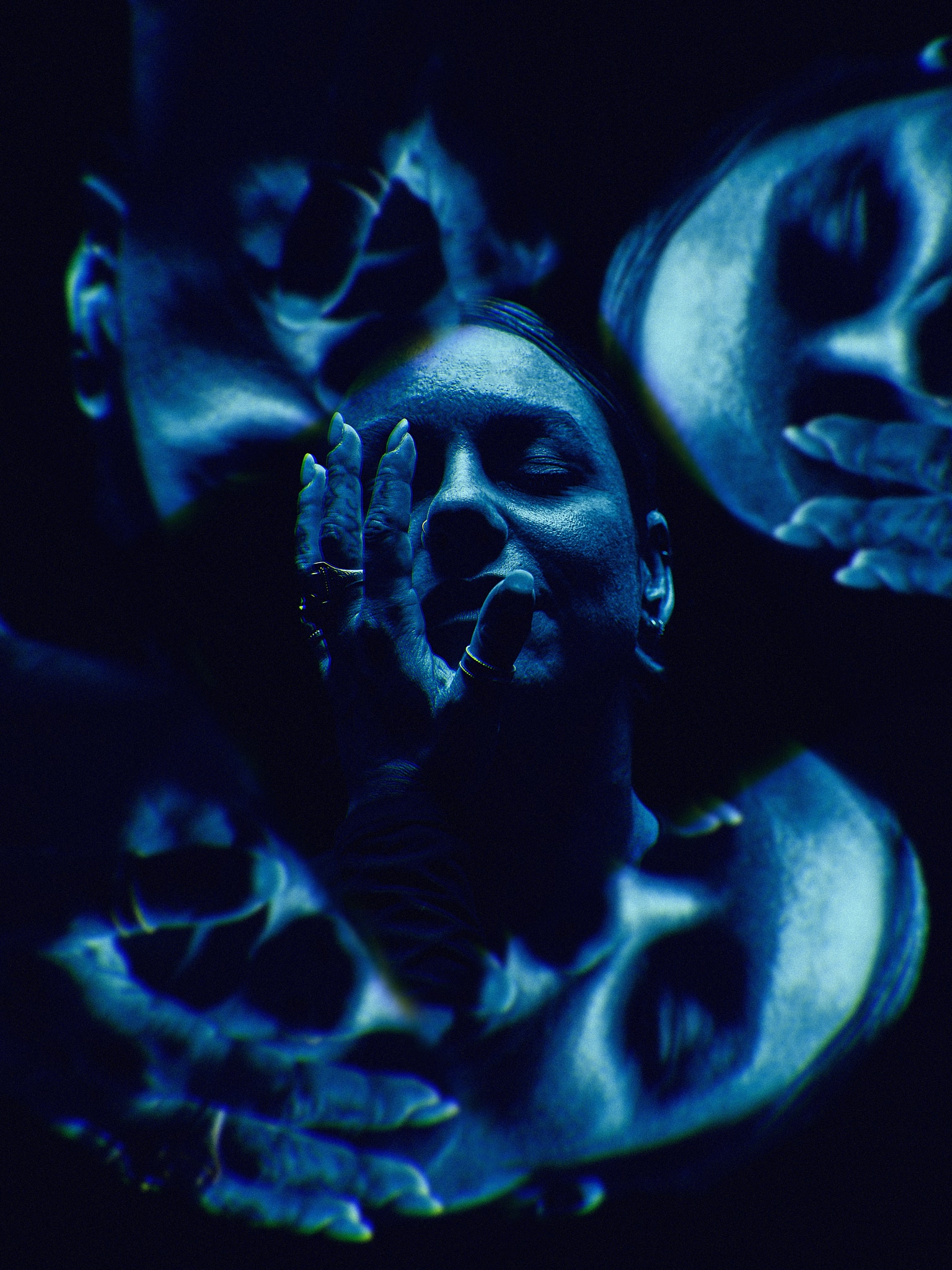 A dark, moody image with multiple overlapping faces of a person, tinted blue. The central figure covers part of their face with their hand while other facial profiles surround them, creating a surreal, dreamlike effect.