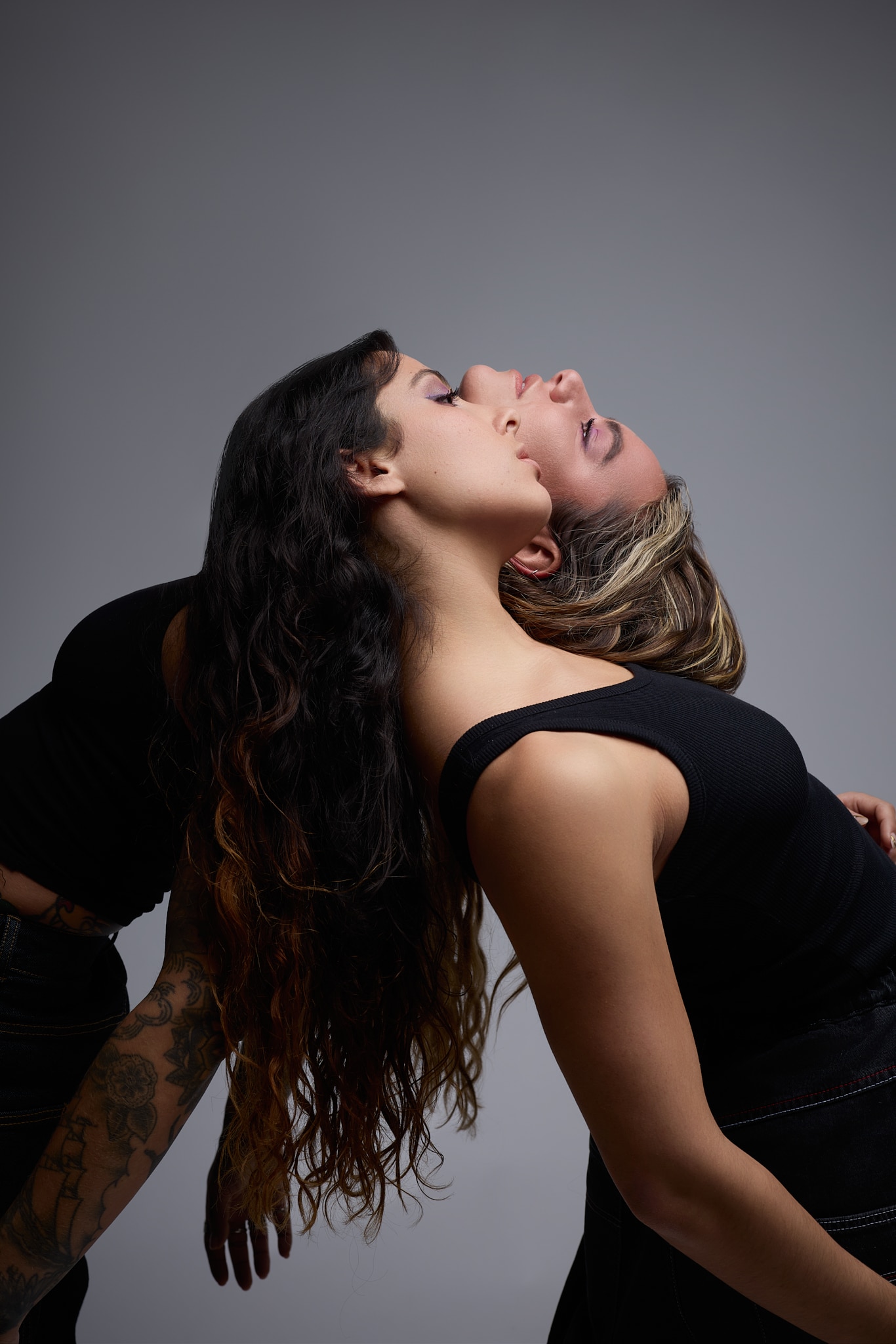 Two women wearing black tops lean against each other, back to back, arching their bodies and tilting their heads back. The background is solid gray, and the tattooed arm of one of the women is visible. They both have long, wavy hair.