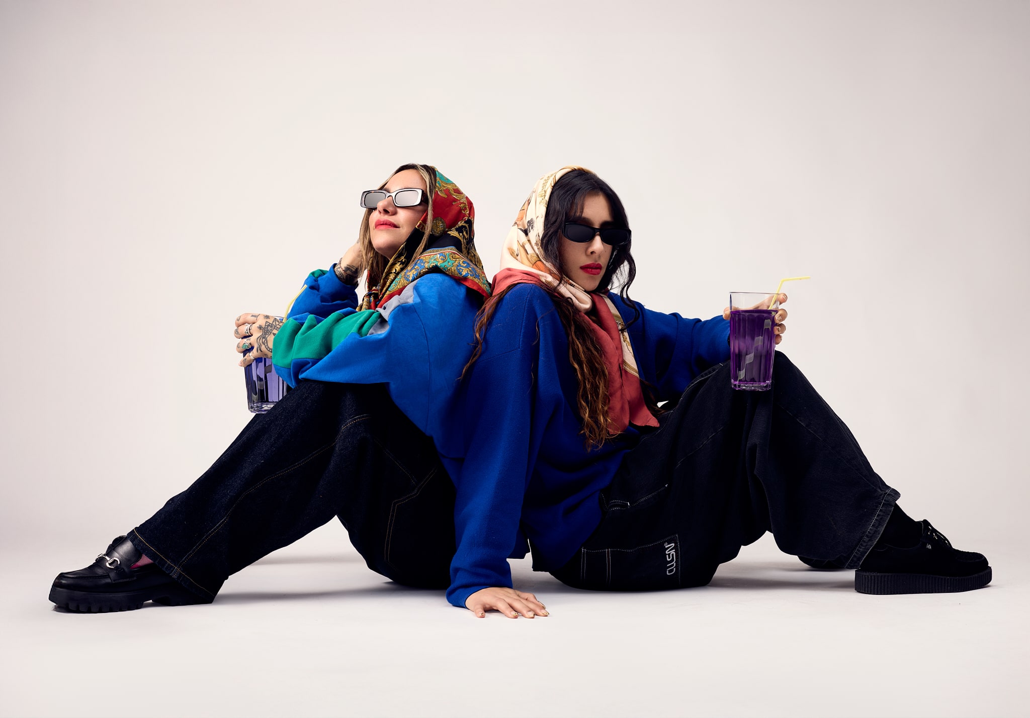Deux femmes portant des foulards colorés, des lunettes de soleil et des sweats à capuche bleus sont assises dos à dos sur le sol, tenant des boissons violettes avec des pailles. L'arrière-plan est uni et de couleur claire.