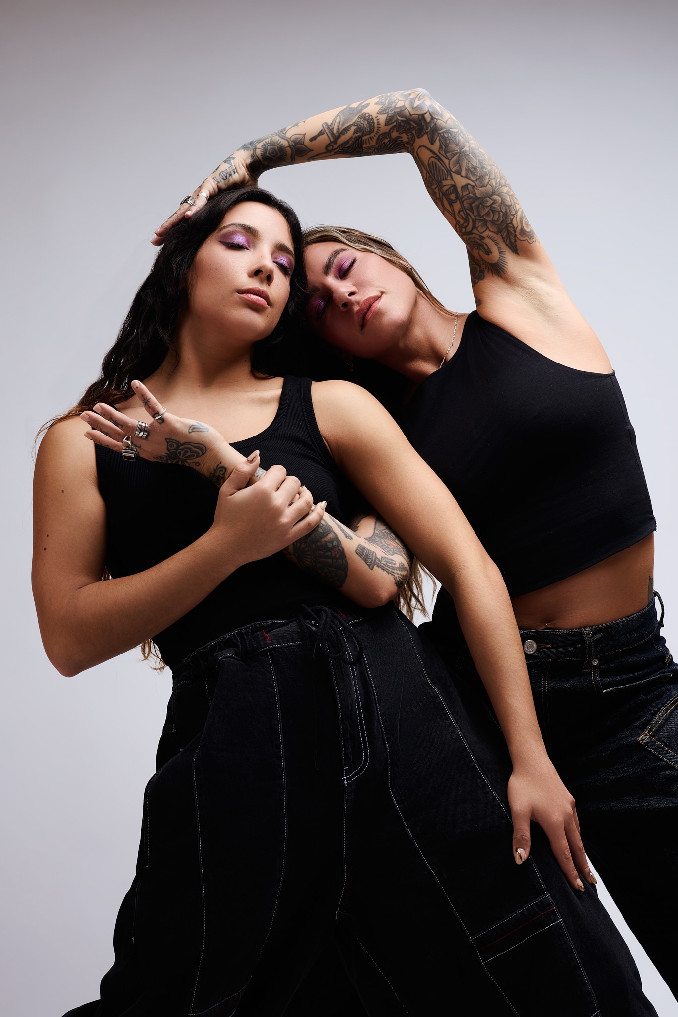 Deux femmes en tenue noire posent l'une près de l'autre. L'une se penche avec un bras au-dessus de sa tête, montrant des bras tatoués, tandis que l'autre croise doucement les bras. Toutes deux ont des cheveux longs et des expressions neutres sur un fond uni.