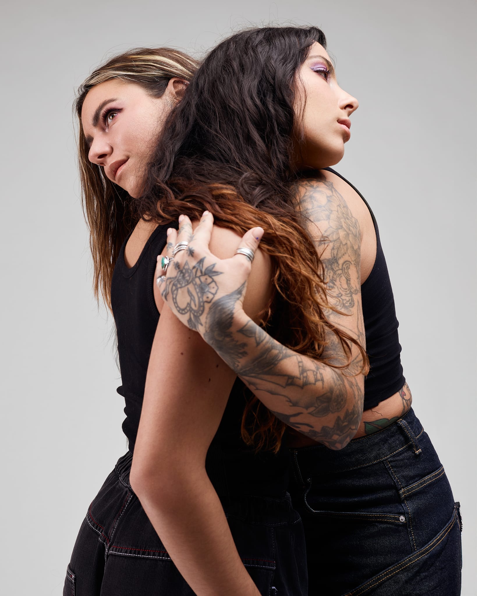Deux personnes aux cheveux longs et tatoués se tiennent dos à dos, s'embrassant doucement. Tous deux portent des hauts noirs et des pantalons sombres, et regardent dans des directions opposées sur un fond clair et uni.