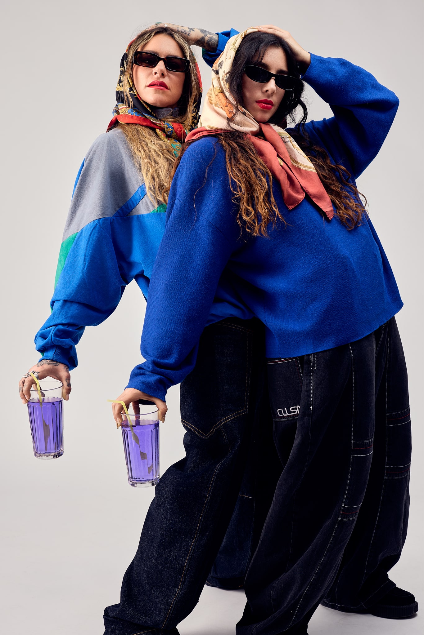 Deux femmes vêtues de pulls bleus, de pantalons sombres et de foulards colorés posent en toute confiance avec des lunettes de soleil et tiennent des verres de liquide violet sur un fond uni.