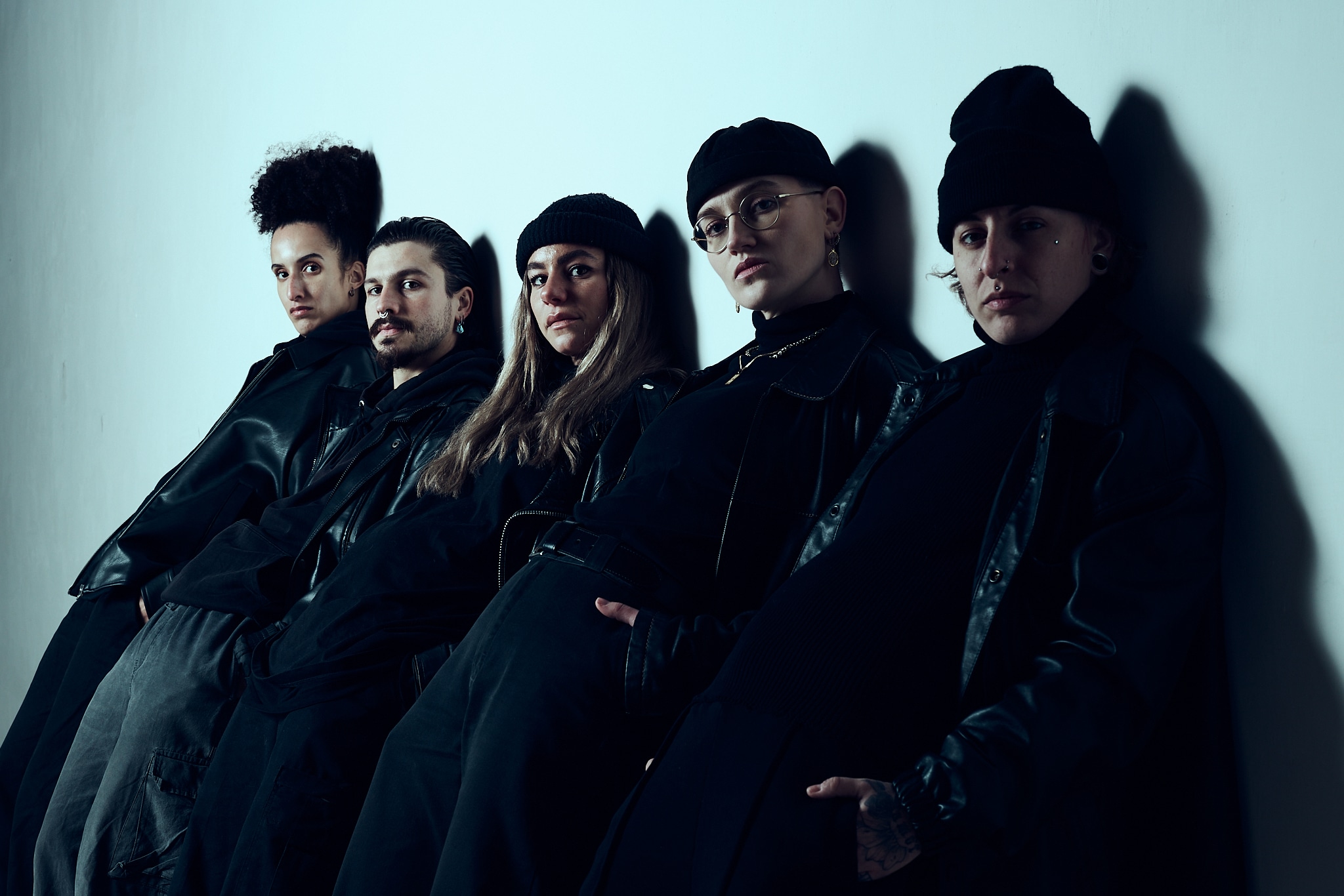 compagnie de danse. Cinq personnes vêtues de vêtements sombres et élégants et de bonnets s'appuient sur un mur blanc, regardant la caméra avec assurance sous un éclairage dramatique et faible.