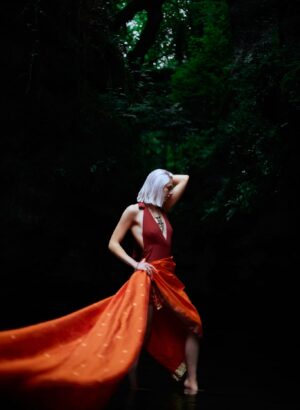 Une personne aux cheveux blancs courts se tient pieds nus dans une eau peu profonde, vêtue d'une tenue rouge et tenant un tissu orange qui coule à propos à côté d'elle, entourée d'un feuillage vert luxuriant dans un cadre naturel faiblement éclairé.