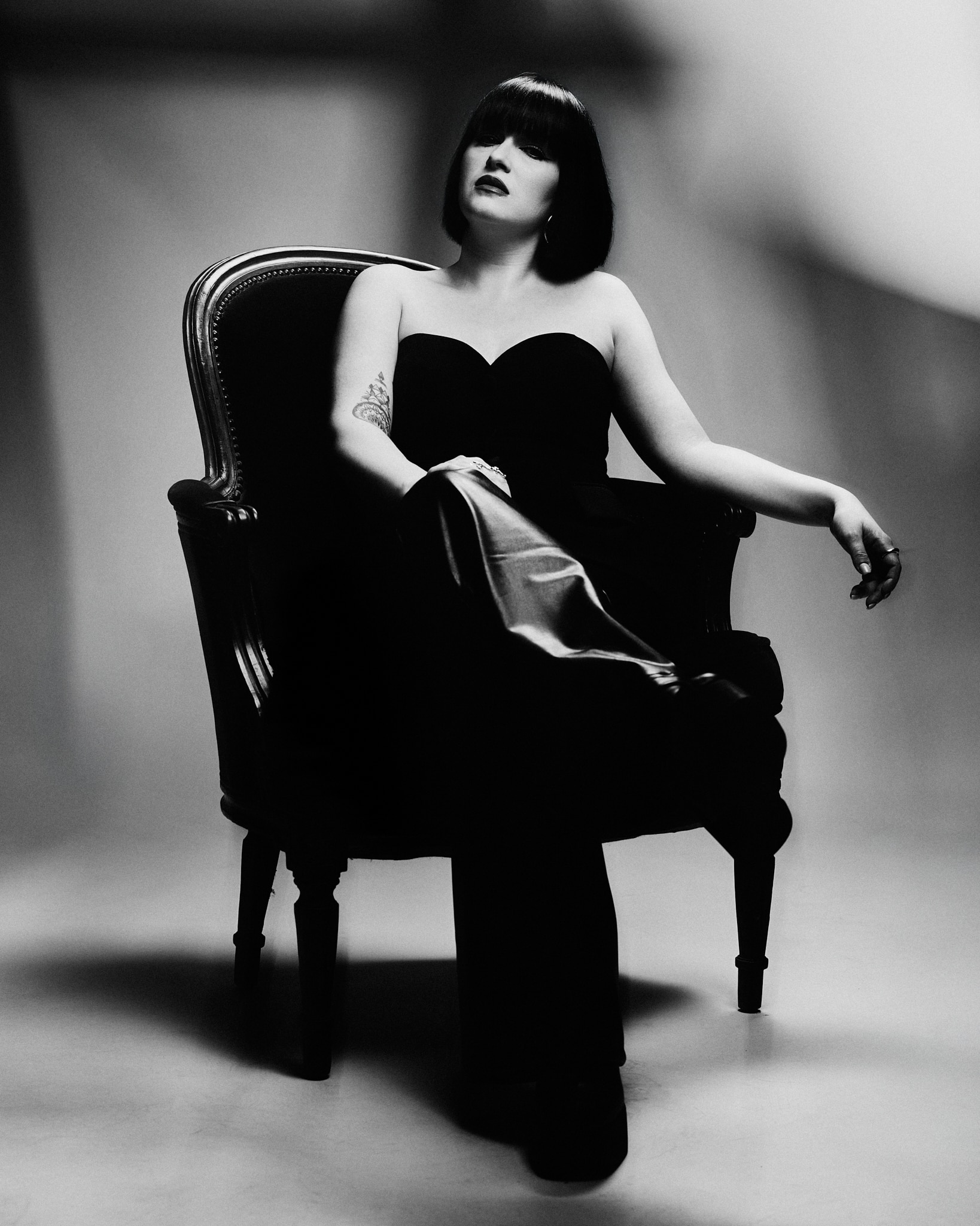 Une femme aux cheveux bruns courts et à la frange est assise avec assurance sur une chaise ornée, vêtue d'une tenue noire sans bretelles. La photo est en noir et blanc et elle porte un tatouage sur le haut du bras. L'arrière-plan est flou.